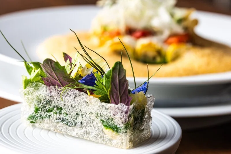 Detailaufnahme eines grünen Salats mit essbaren Blüten auf einem weißen Teller, im Hintergrund ein Gericht mit Pasta und Gemüsen.