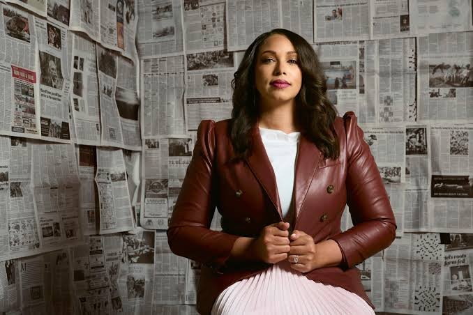 A woman with dark hair wearing a burgundy leather jacket sits in front of newspaper-covered walls.