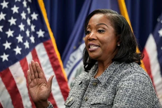 A woman with dark hair taking an oath or speaking, with her right hand raised, in front of American flags and a blue curtain.