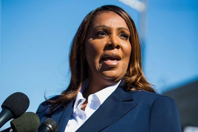 A woman wearing a navy blazer and white shirt speaking into multiple microphones outdoors with a clear blue sky in the background.