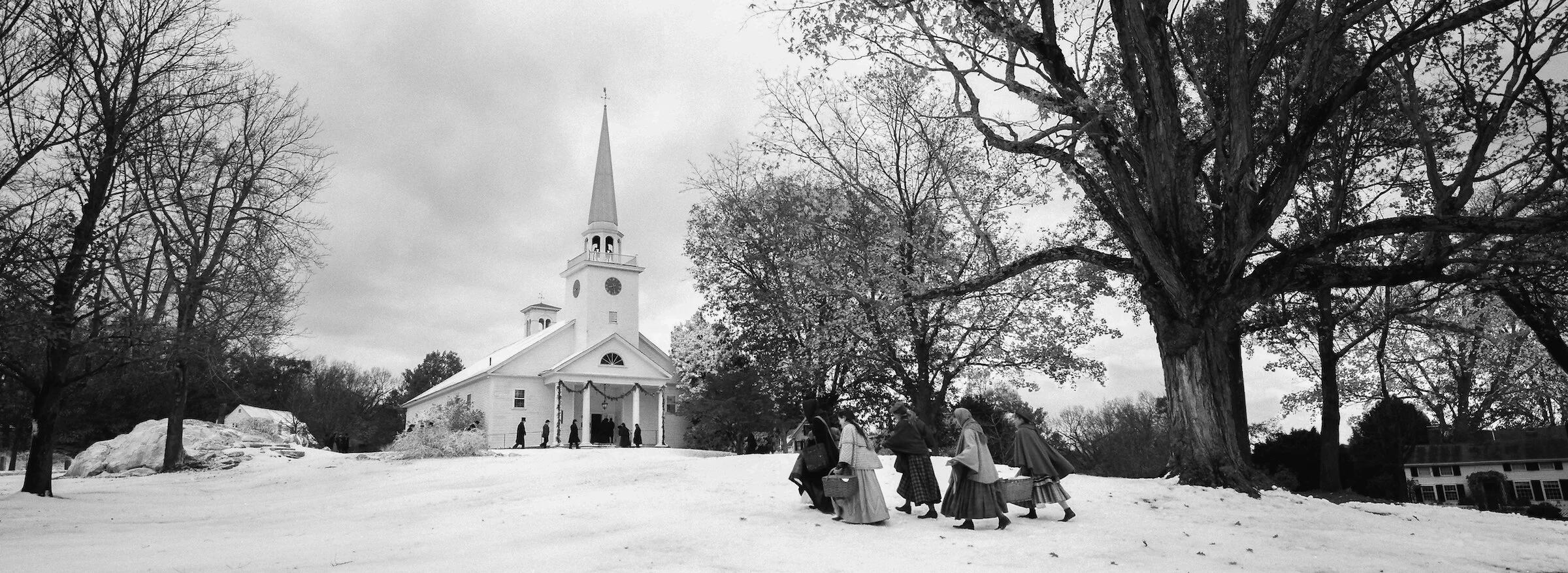 A black and white photo of a church with a tall steeple, surrounded by leafless trees. Several people dressed in historical clothing are walking towards the church, carrying baskets and belongings.