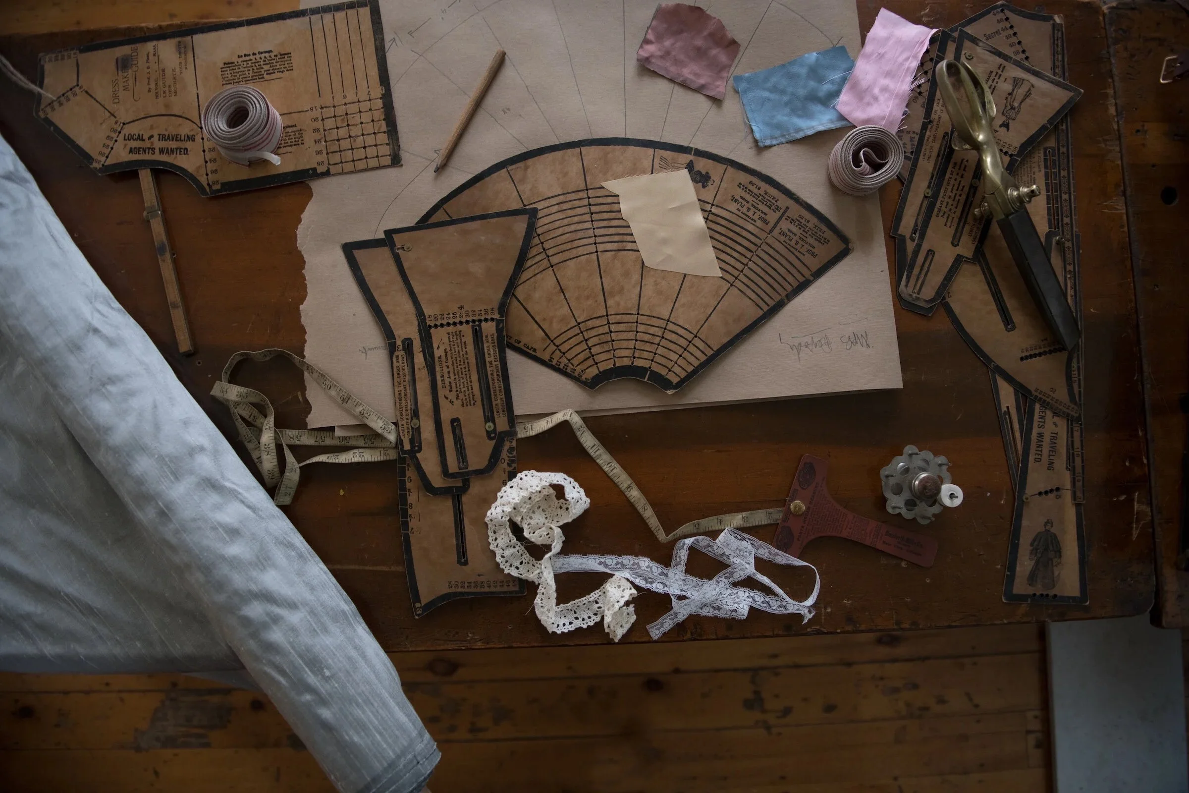 A sewing or pattern-making workspace with vintage dress pattern pieces, fabric swatches, measuring tape, a vintage seam roller, and lace trim on a wooden table.
