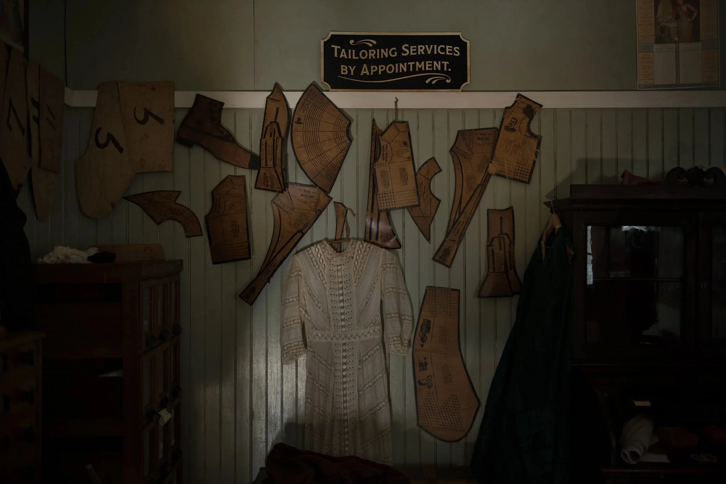 Display of fabric patterns and tailoring materials on a wall, with a sign that reads 'Tailoring Services by Appointment.' and a vintage white dress hanging in the center.