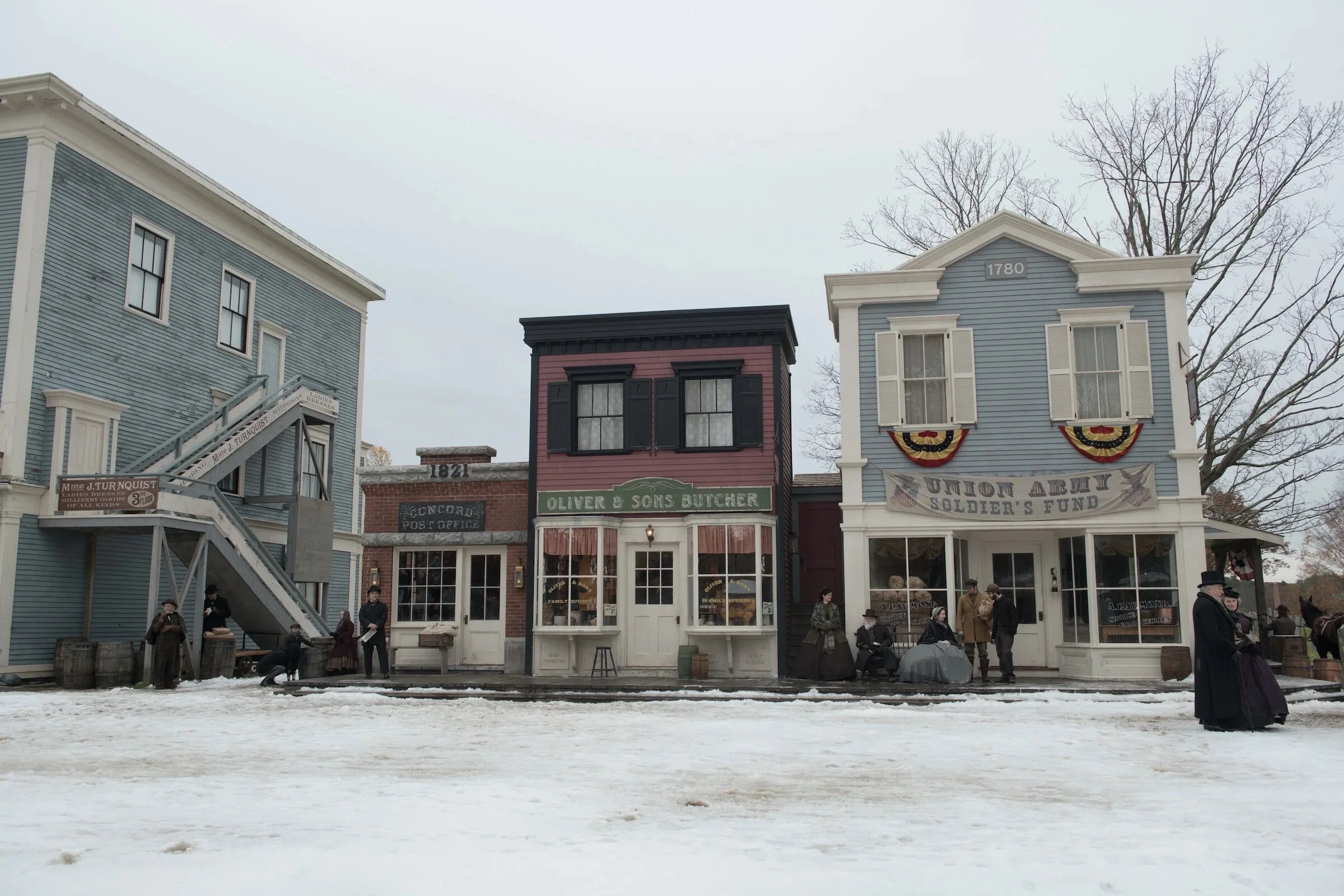 Historic buildings with vintage storefronts and people dressed in period clothing, suggesting a historical reenactment or old-town setting on a snowy day.