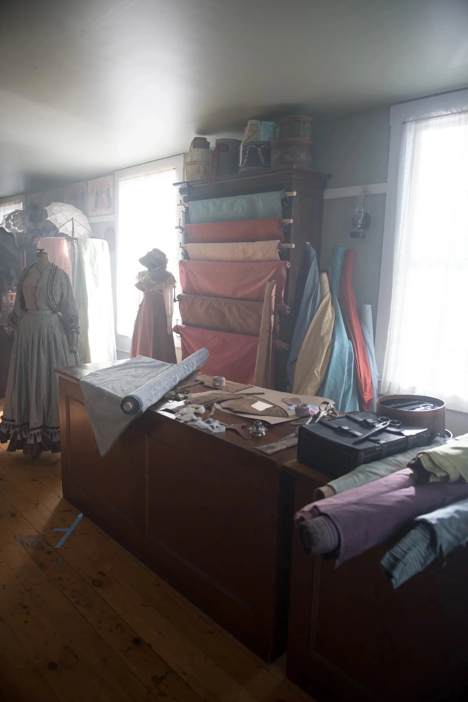A craft room with mannequins dressed in vintage clothing, a table with fabric and sewing supplies, and a wall of fabric bolts in various colors. Natural light streams through the windows.