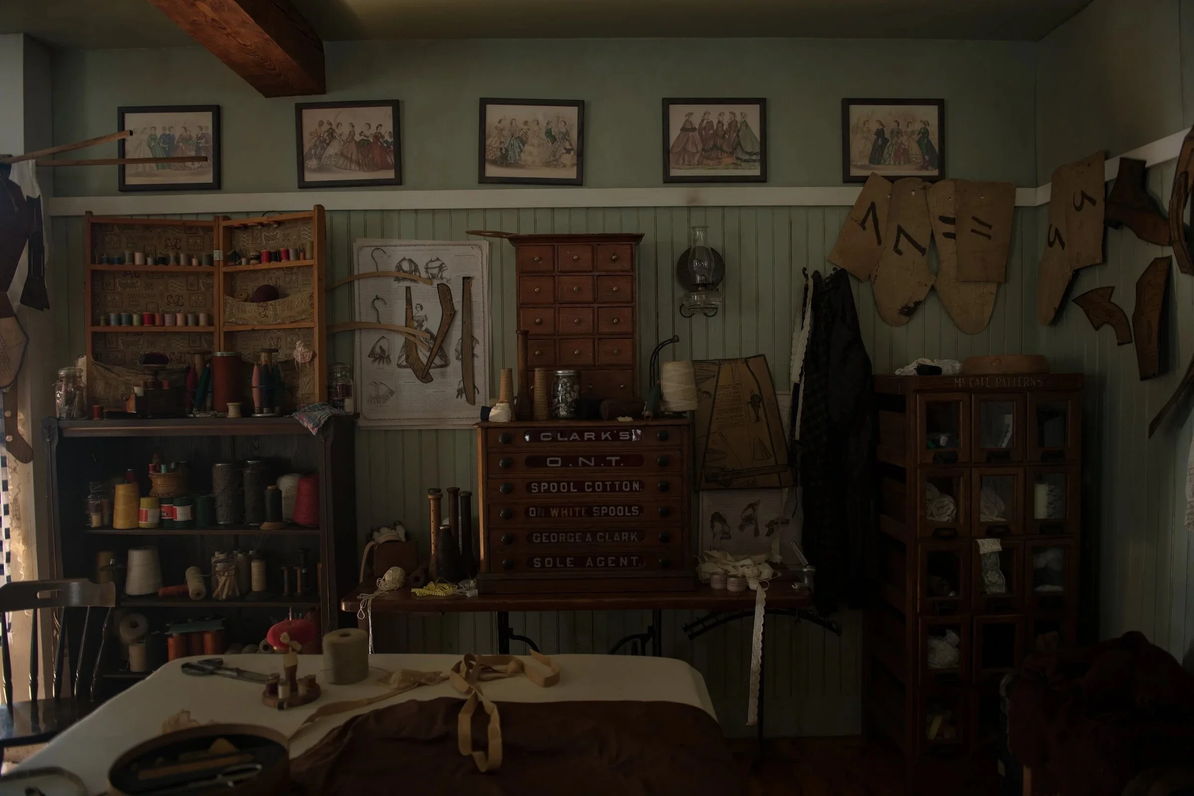 Craft room with shelves of sewing supplies, framed historical fashion illustrations on the wall, a wooden chest of drawers, and fabric patterns hanging on the wall.
