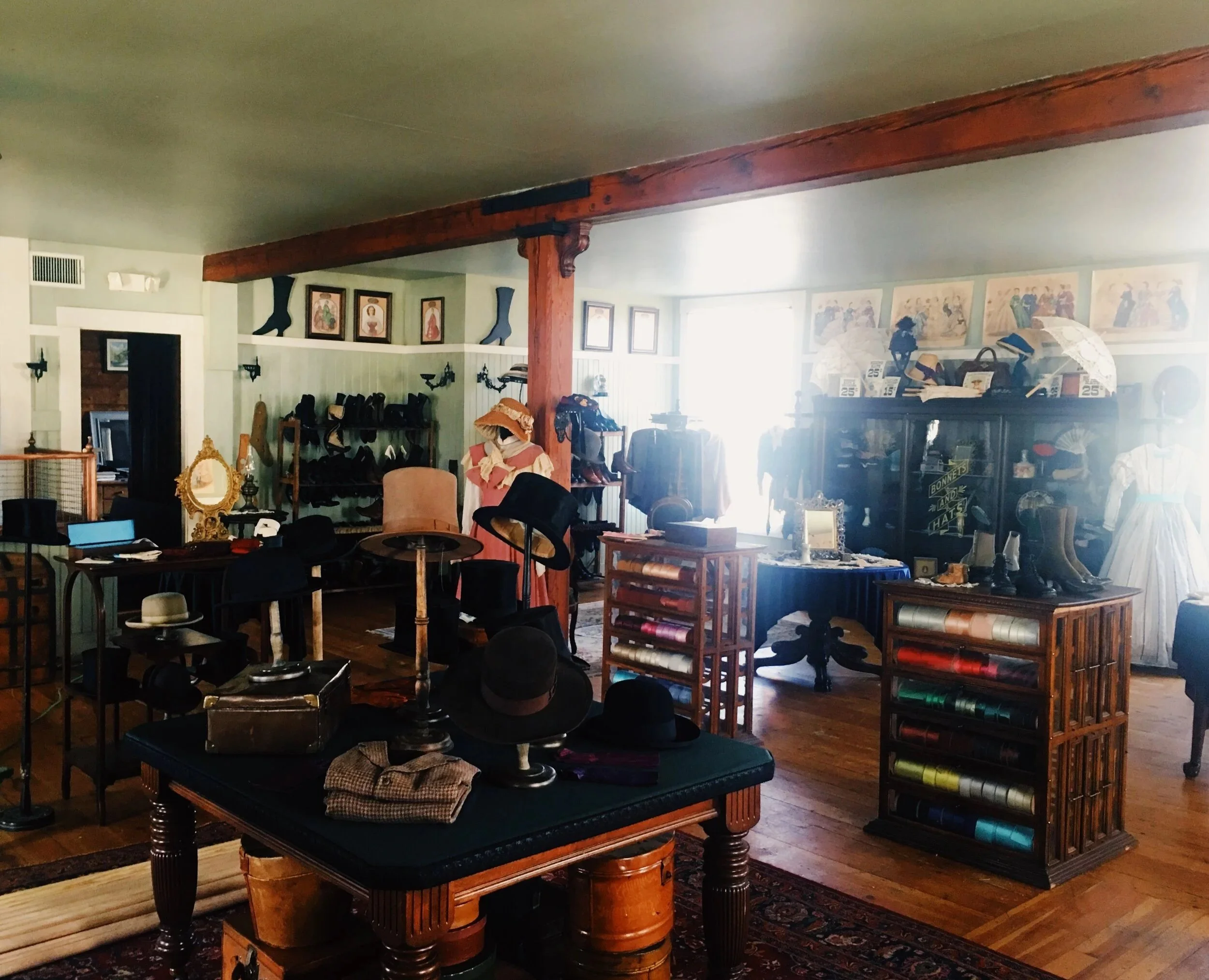 A vintage store interior with various hats, shoes, picture frames, and furniture, including wooden tables and bookshelves, with floral paintings and decorative items displayed.