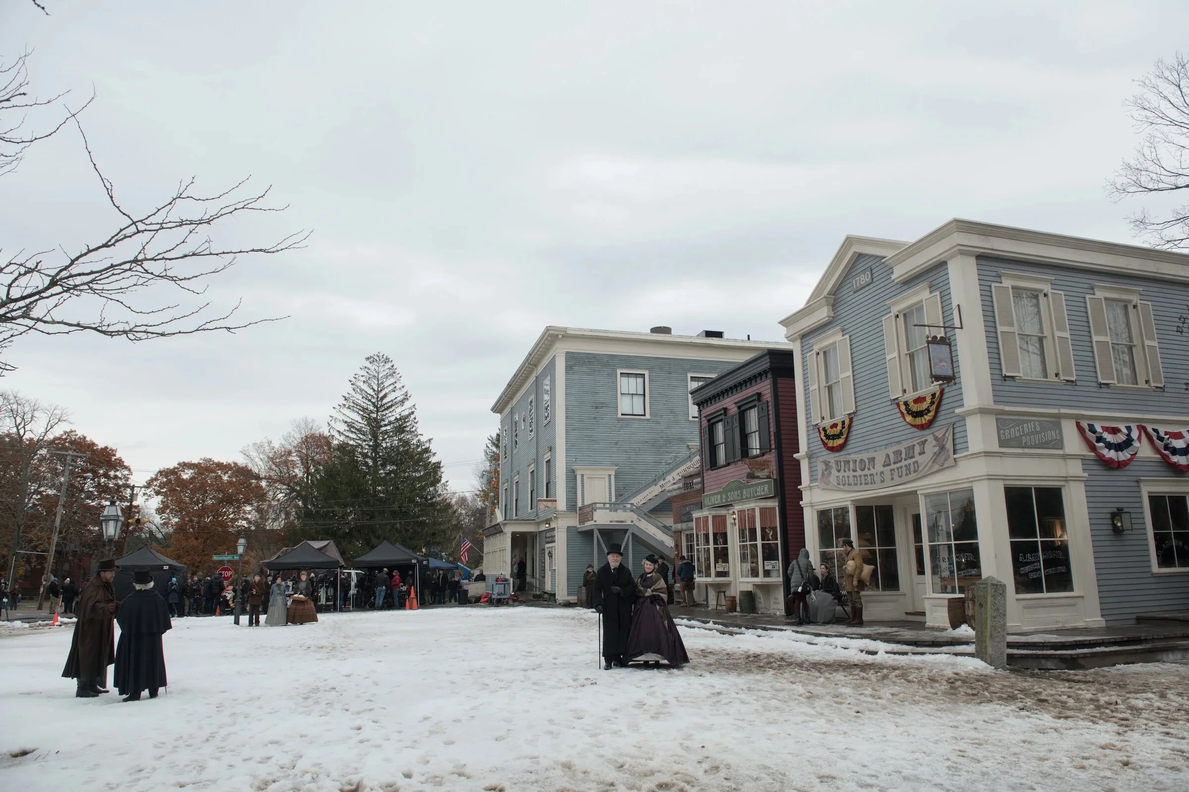 Historic small-town street scene with colorful buildings, people dressed in period costumes, and snow on the ground, suggesting a reenactment or patriotic event.