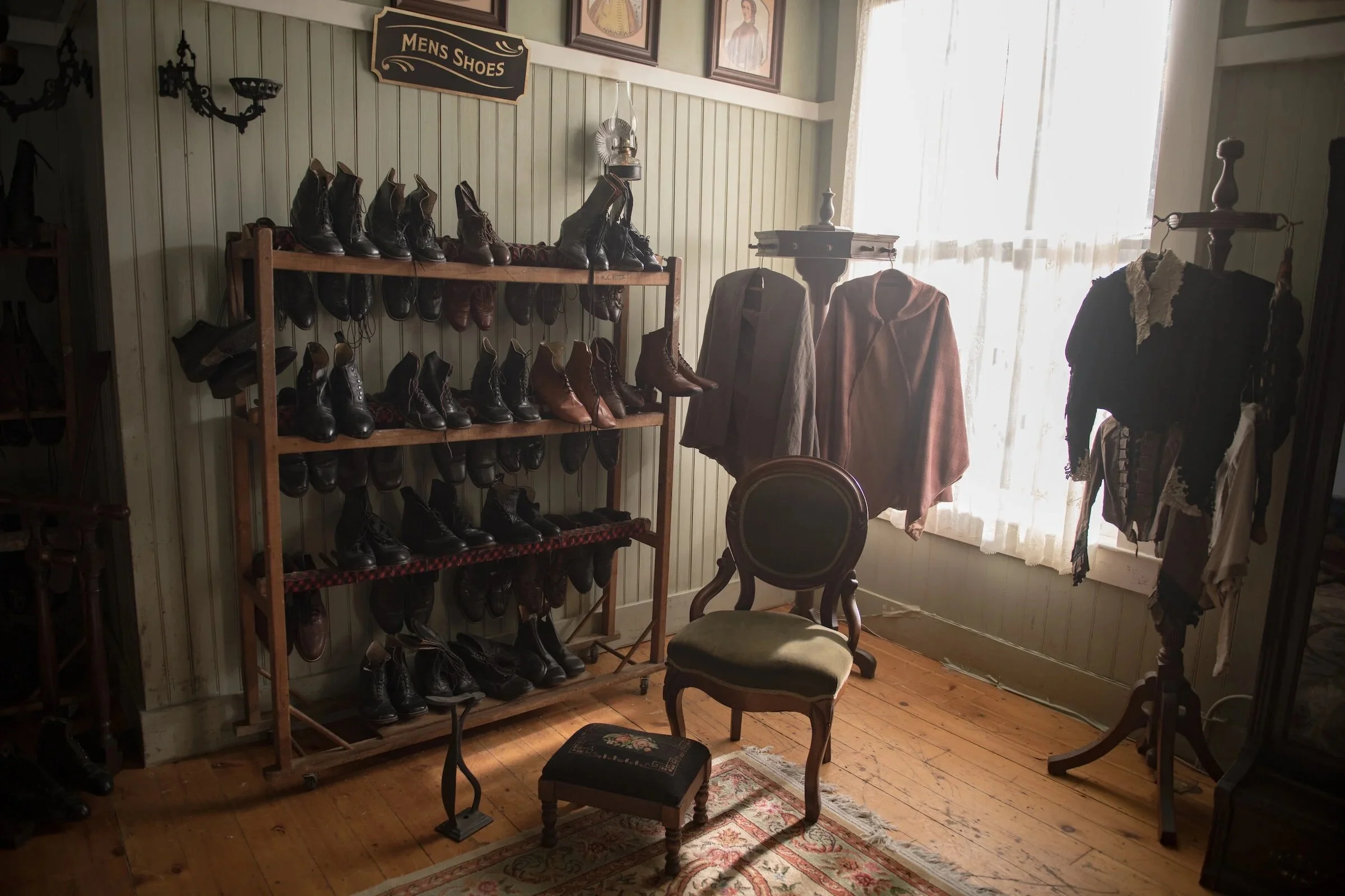 Photo of a vintage shoe store showing a wooden shelf filled with men's leather shoes and boots. There is a sign above the shelf that says 'Men's Shoes,' and the store has wooden floors, a floral rug, chairs, and a window with sheer curtains allowing natural light.