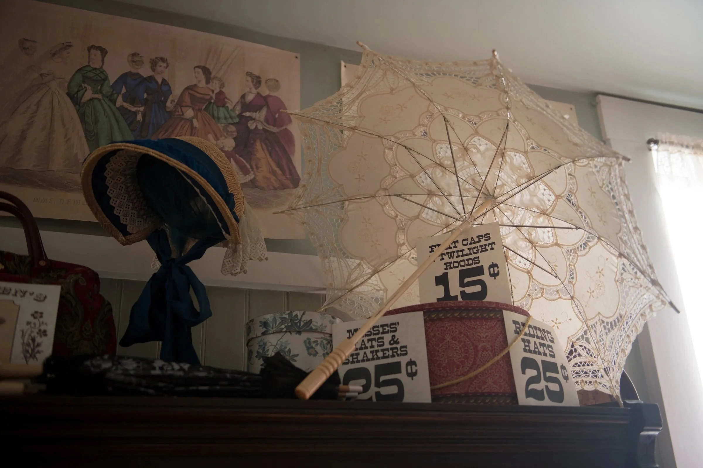 A display of vintage items including lace umbrellas, hats, and a vintage fashion illustration on the wall with a sign for sale prices.