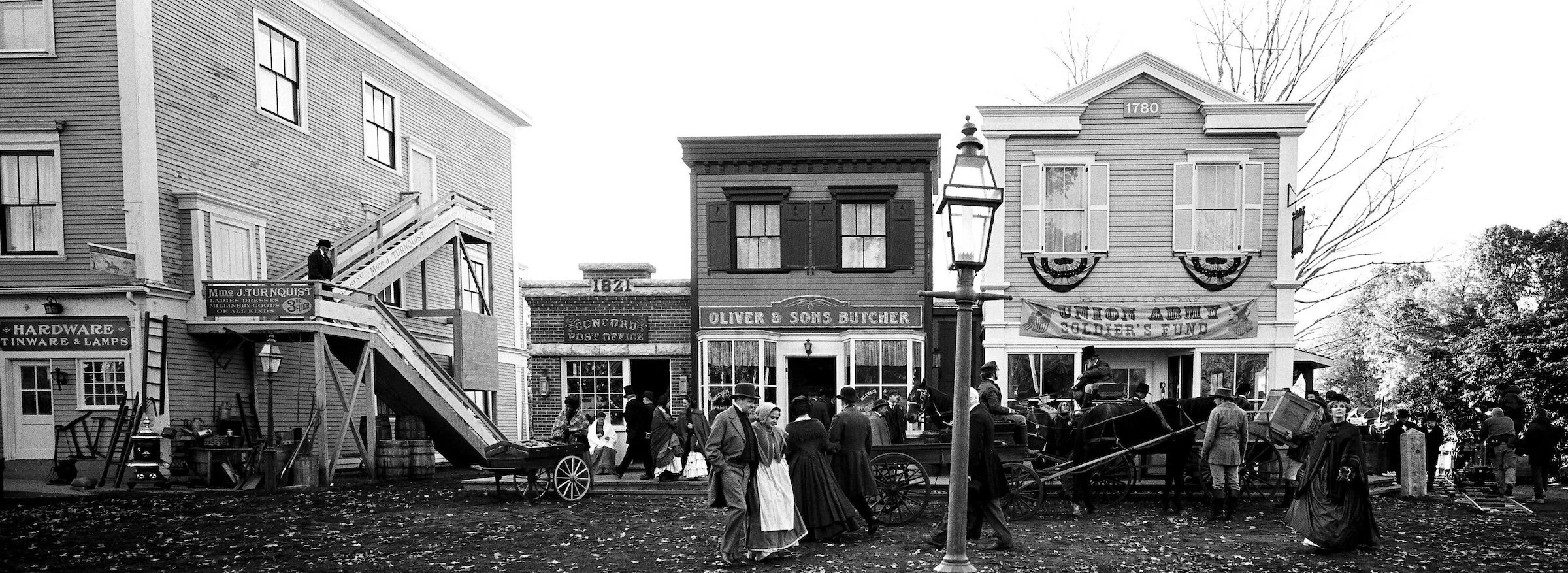 Historical reenactment scene on a street with people dressed in period clothing and horse-drawn carriages, stores with vintage signs, and a banner supporting soldiers' fund.