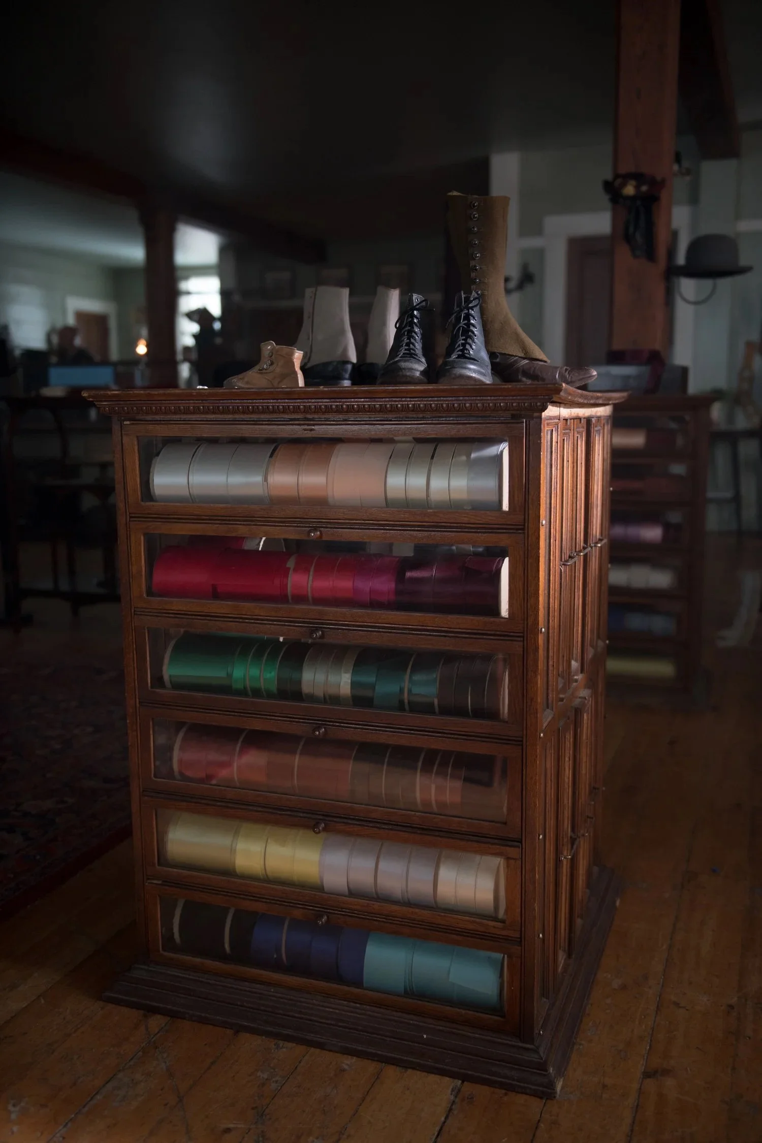 A wooden display case with multiple drawers containing color swatches, topped with vintage boots and shoes, in a room with wooden floors and furniture.