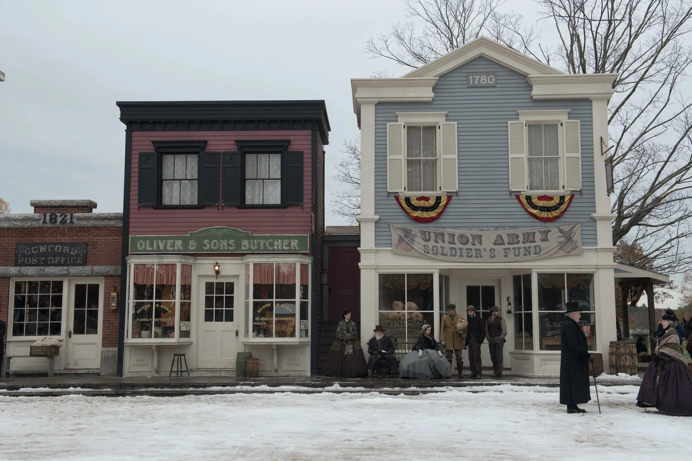 A historical reenactment scene with 19th-century buildings, including a butcher shop and a post office, with people dressed in period attire and gathered in front of the buildings.