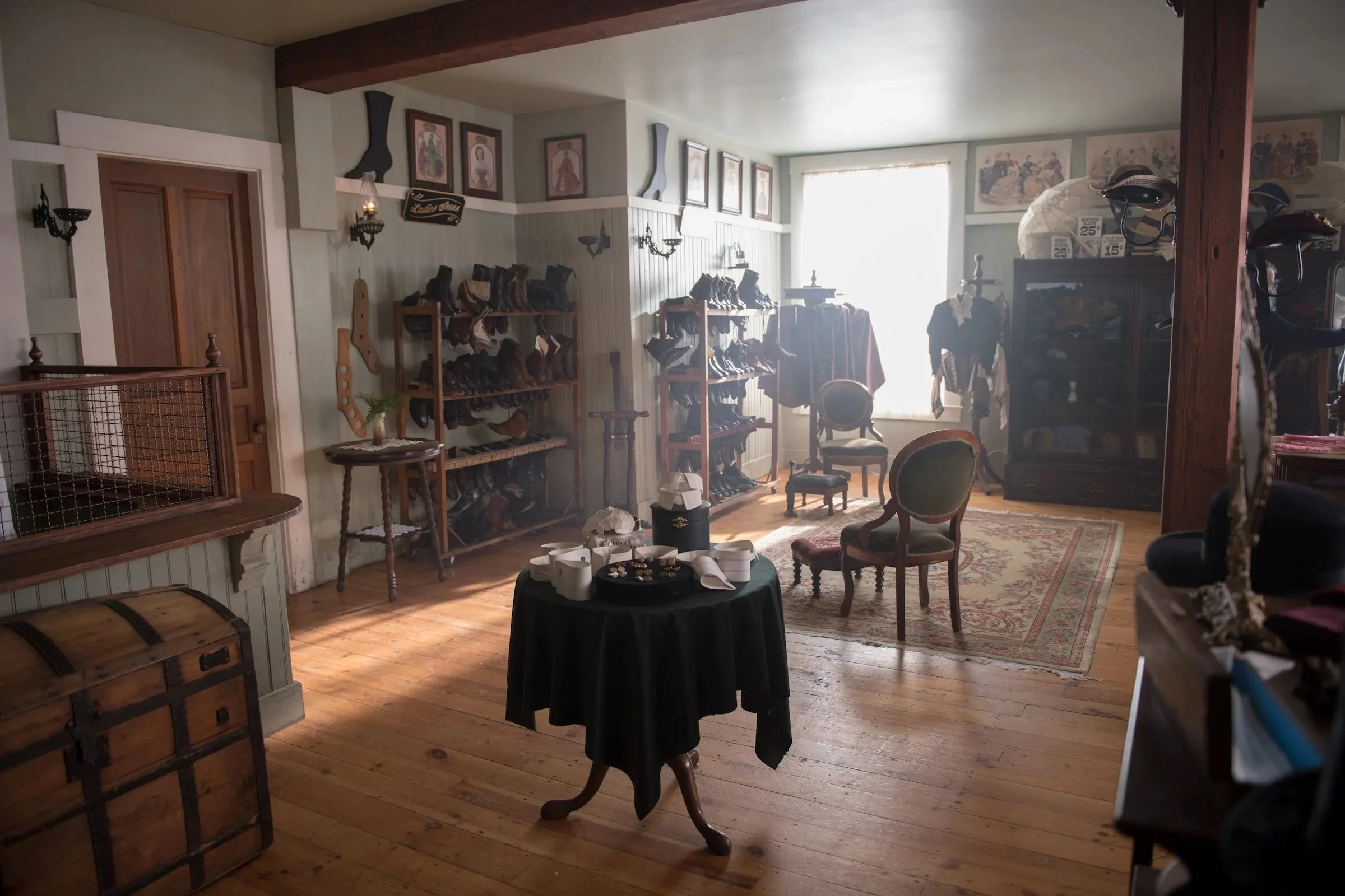 A room filled with vintage items including shelves with shoes, a wooden chest, and framed artwork on the walls. There is a table with jewelry and a small potted plant, and chairs with decorative upholstery. Natural light comes through a large window, illuminating the space.