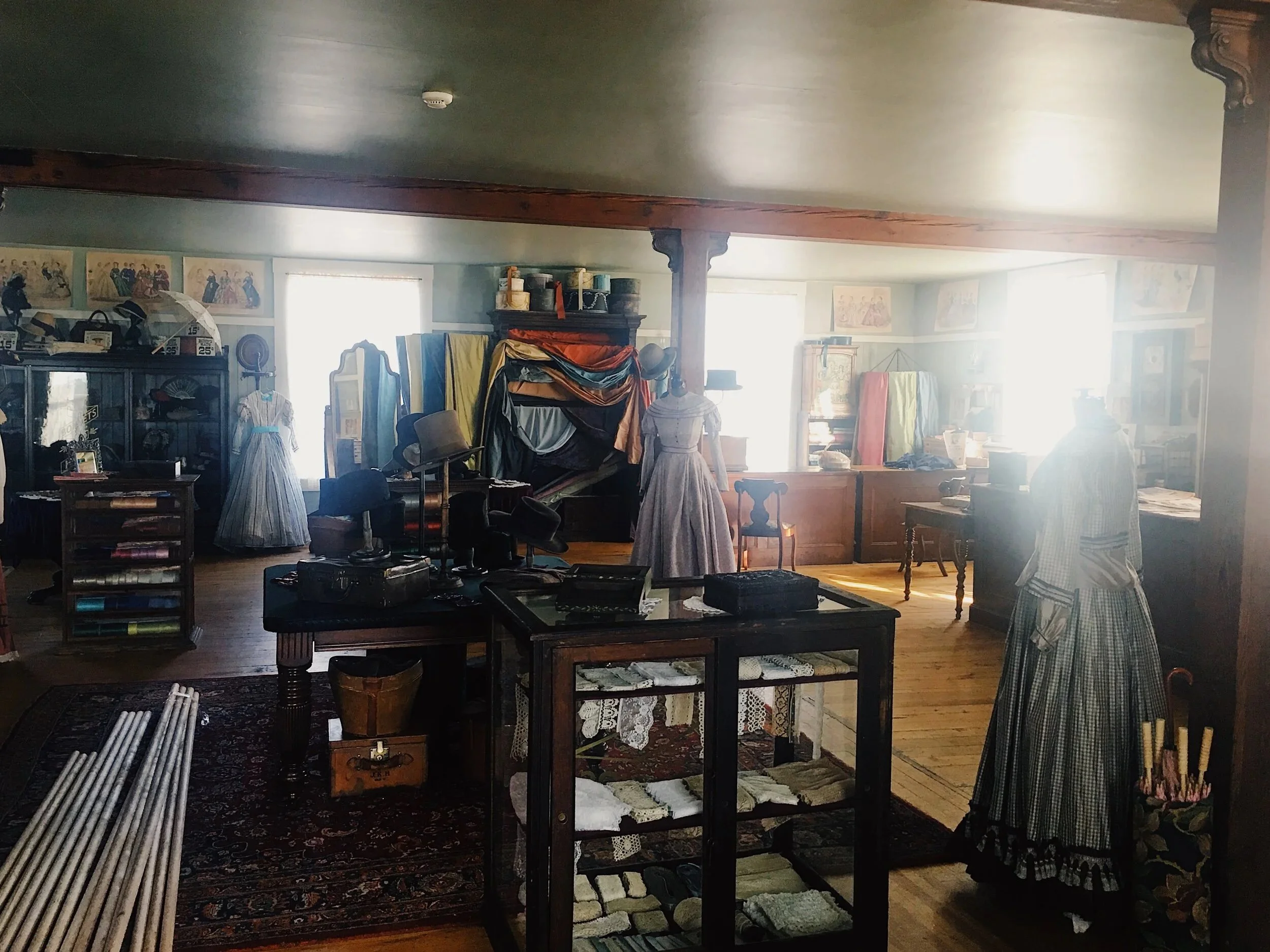 Interior of a vintage store filled with textiles, dresses on mannequins, shelves with fabric rolls, framed artwork on the walls, and various decorative objects, illuminated by natural light from large windows.