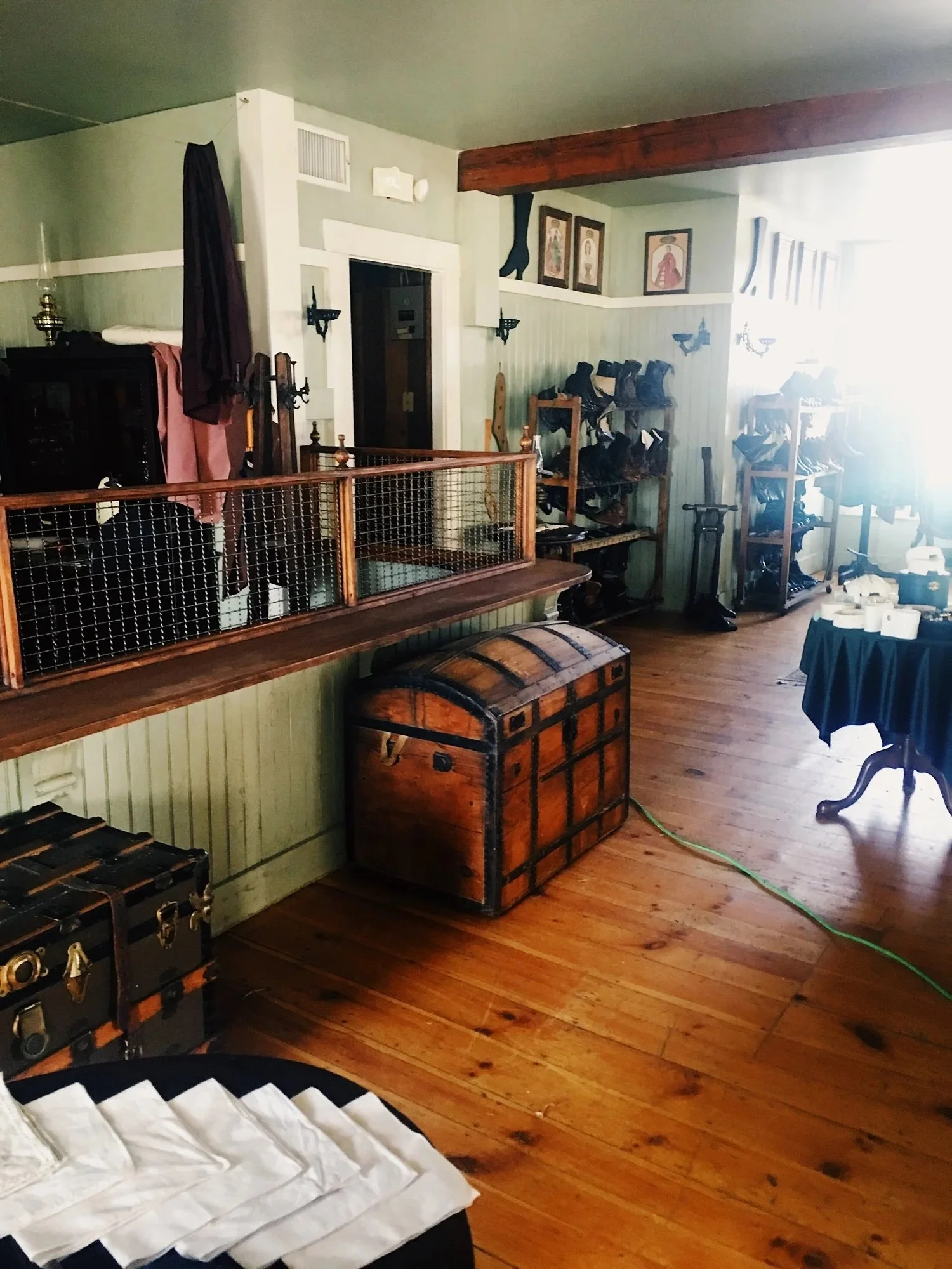 A cozy room with wooden flooring, green walls, and various vintage furniture and decorations, including a wooden chest, shoe racks, a table with rolls of paper, and framed artwork on the wall.