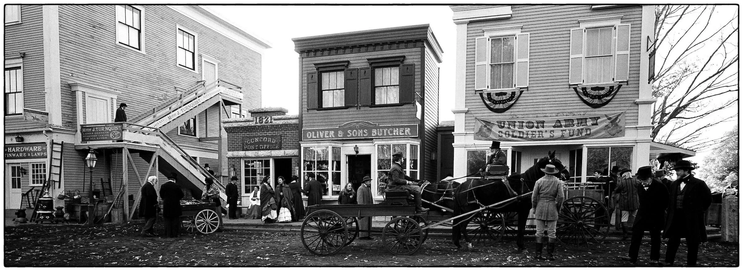 Historical black-and-white scene of a street with people, horse-drawn carriages, and buildings with signs, resembling an old colonial town.