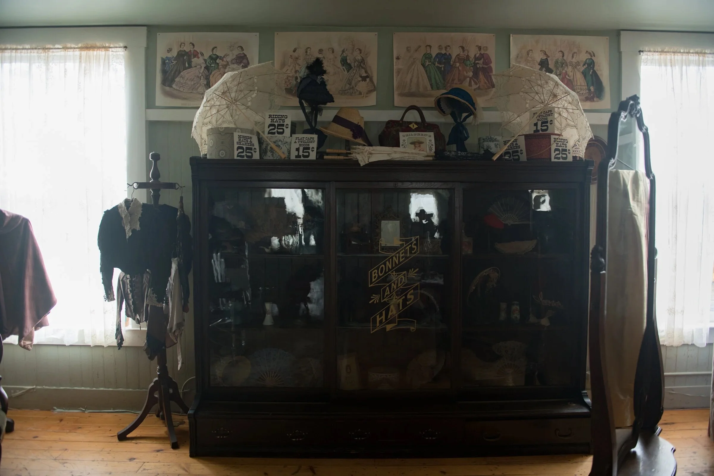 A vintage display cabinet filled with old hats, parasols, and accessories, with framed Victorian-style artwork hanging on the wall behind it.