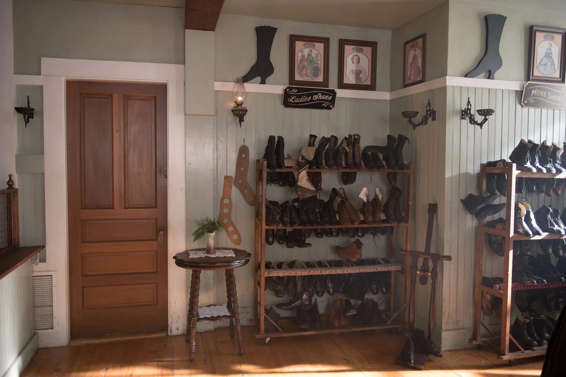 A wall with framed vintage fashion illustrations and decorative shoe silhouettes, wooden shoe racks filled with various pairs of boots and shoes, a small round wooden table with a lace doily and a potted plant, and signs labeled 'Ladies Shoes' and 'Men's Shoes'.