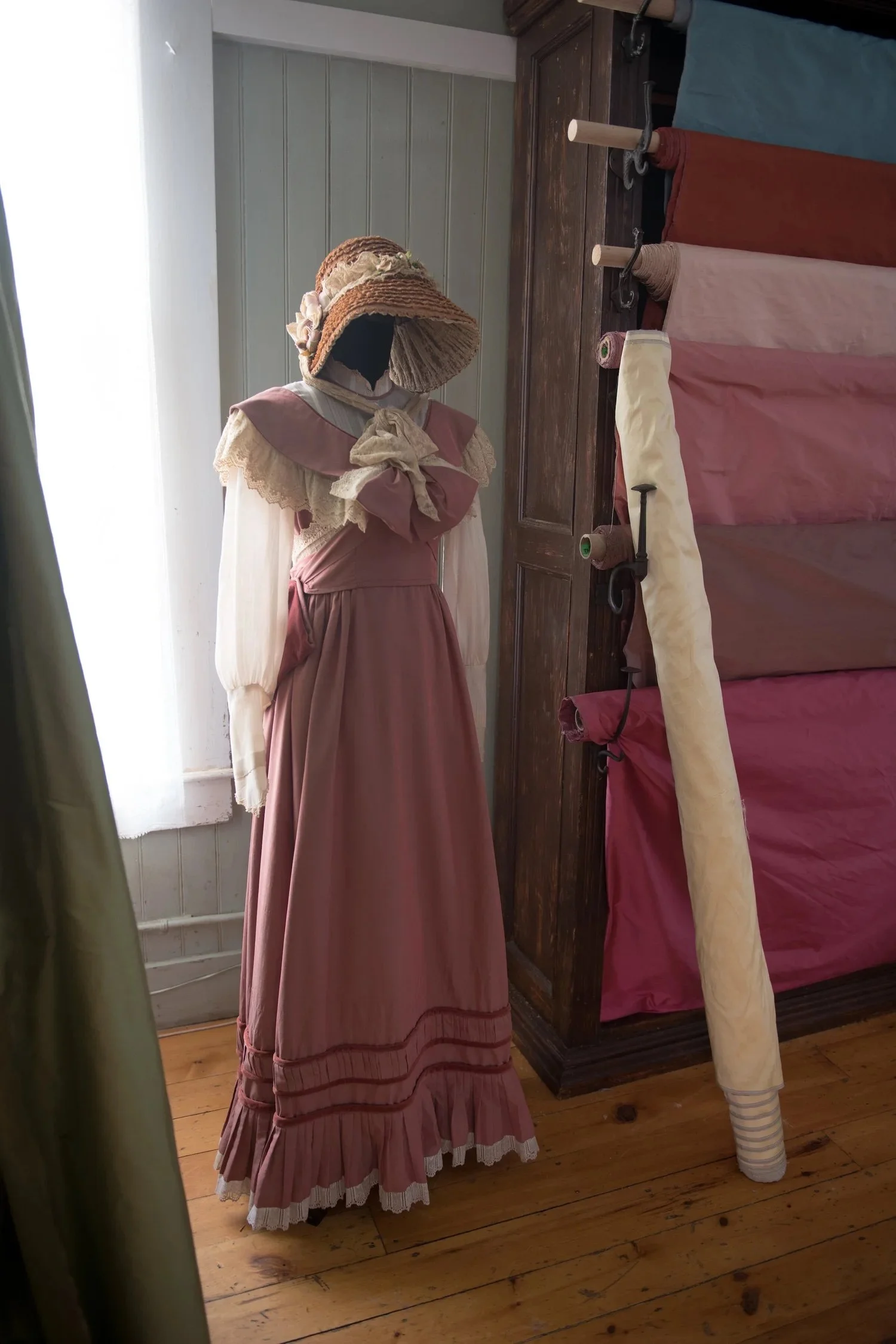 A vintage-style dress with long sleeves, lace details, and a large bow at the neck, displayed on mannequin. The dress is in shades of pink and cream. The mannequin is wearing a wide-brimmed straw hat decorated with fabric flowers.