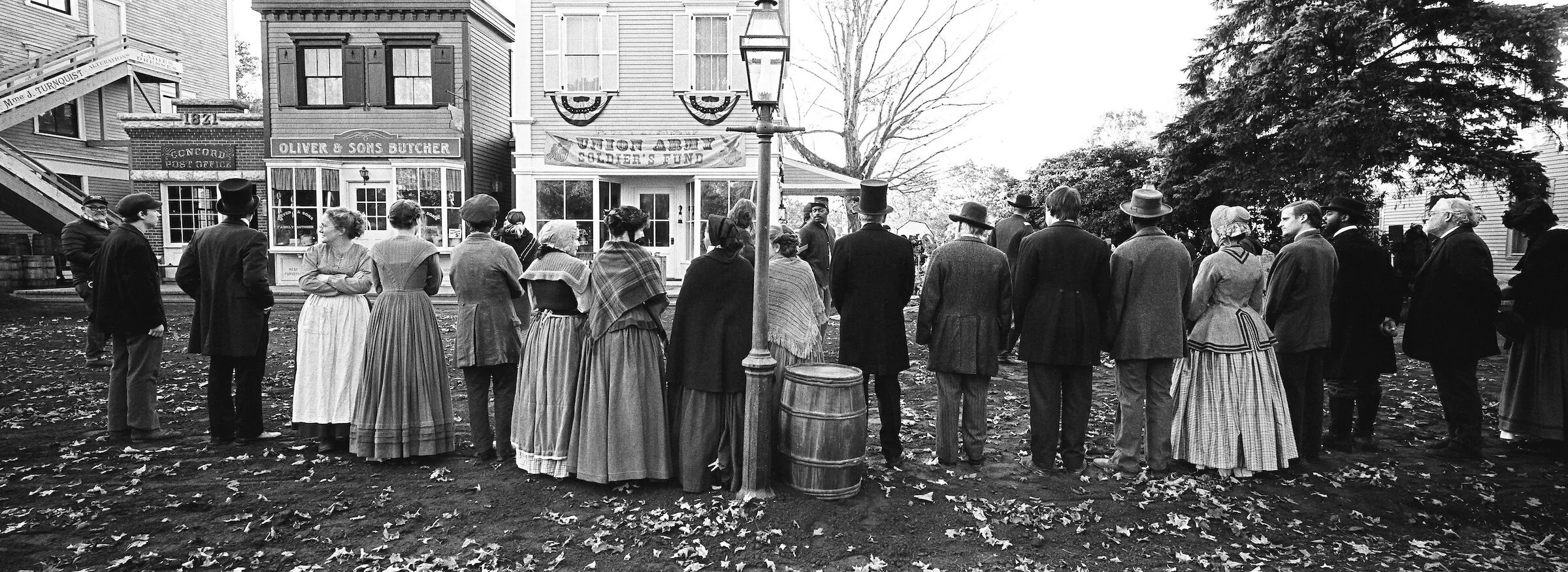 A black and white photo of people dressed in 19th-century clothing standing in a line outdoors, possibly for a historical reenactment or event, with buildings and trees in the background.