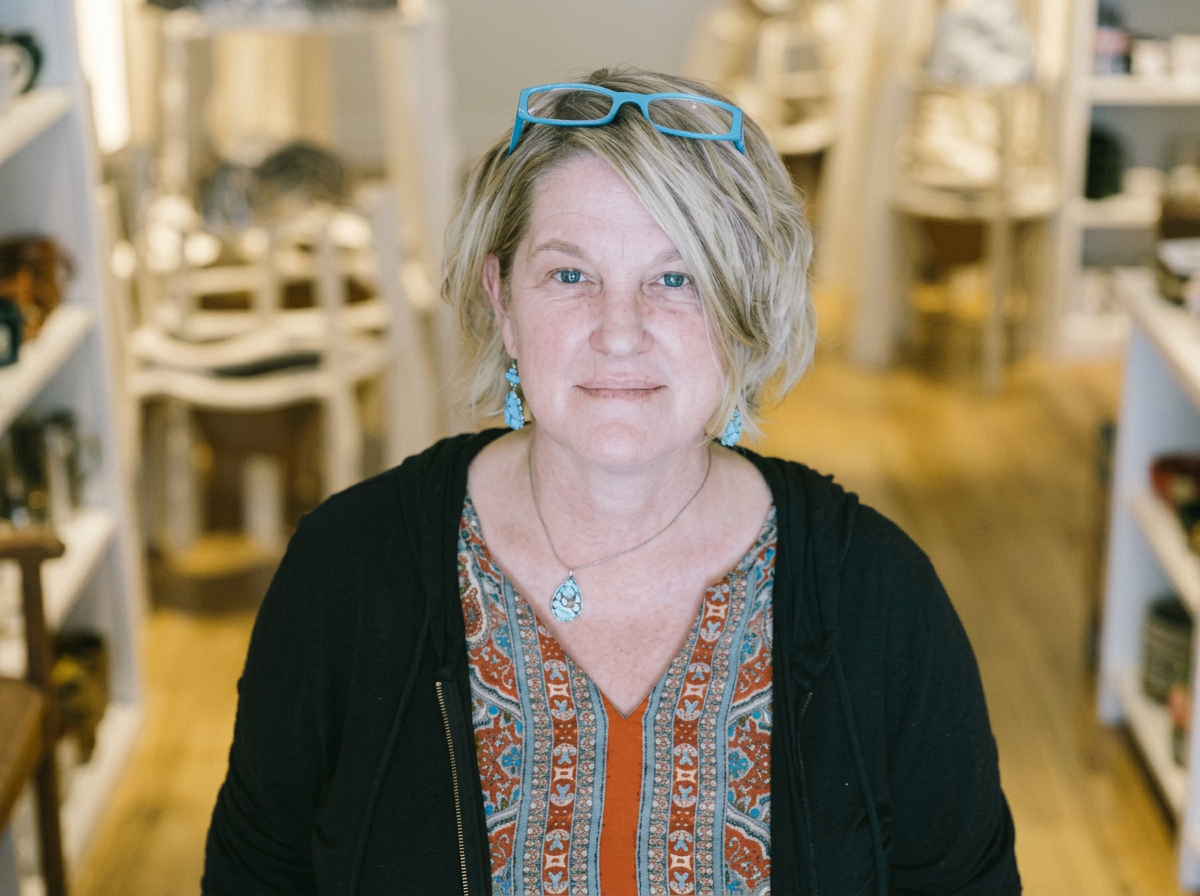 A middle-aged woman with short blonde hair, wearing blue glasses on her head, a black jacket, turquoise jewelry, and an orange patterned blouse, standing in a store with white shelves and wooden flooring.
