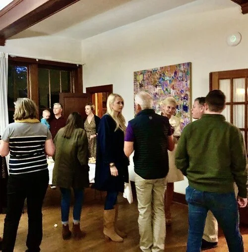 Group of people socializing in a warmly lit room with wooden floors and a colorful abstract painting on the wall.