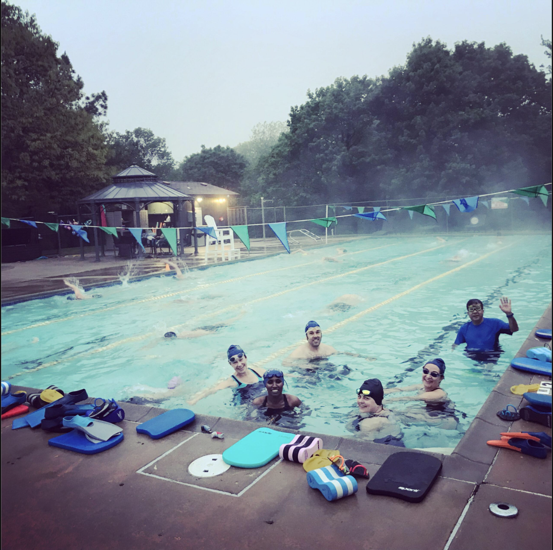 Group of people enjoying a swim in an outdoor pool with swim gear and poolside equipment in the foreground, trees in the background, and decorative flags across the pool.