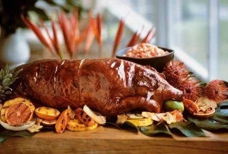 Roast pig with vegetables and a bowl of rice in the background on a wooden table.
