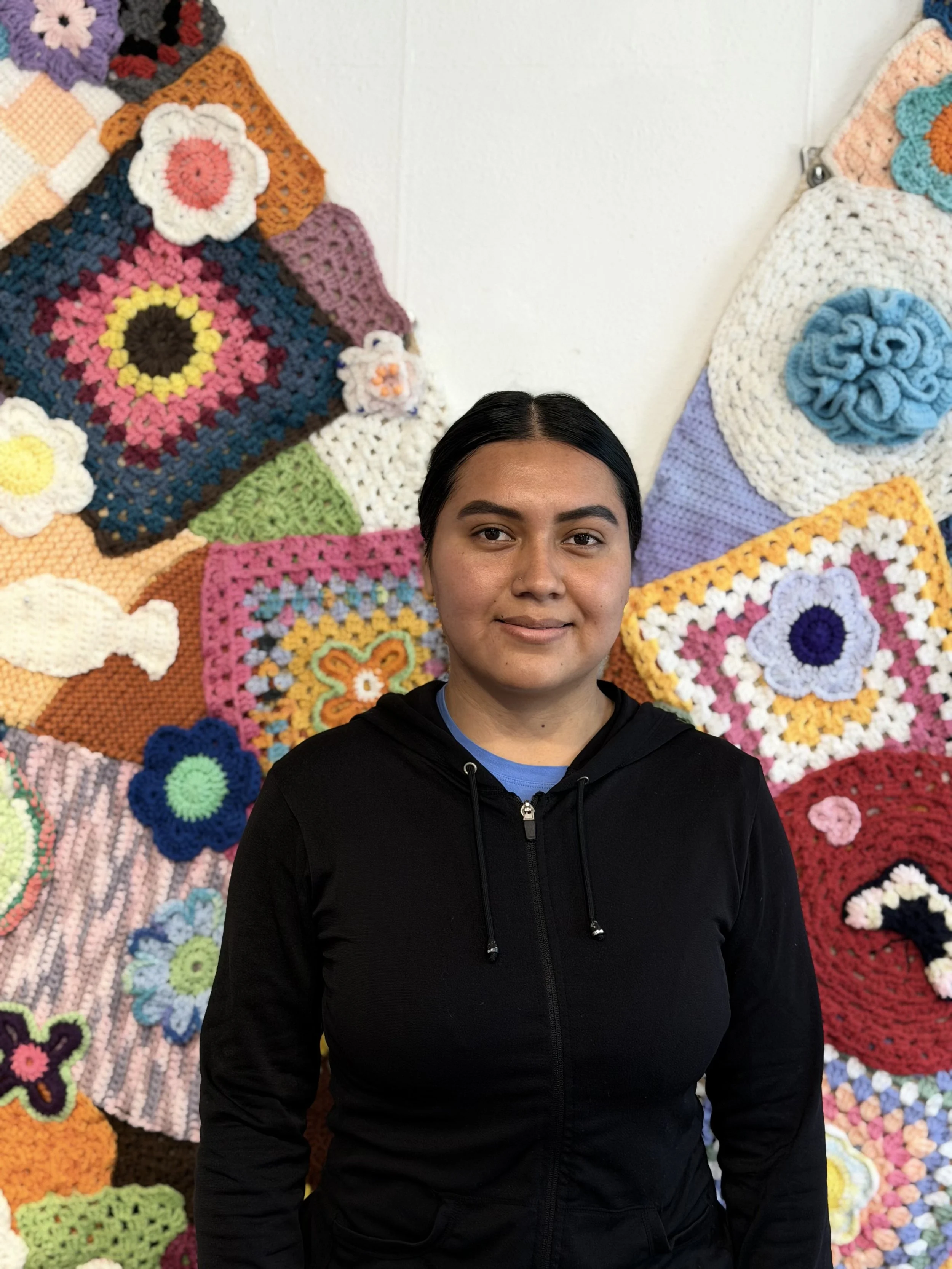 A woman with dark hair, wearing a black hoodie, standing in front of a wall decorated with colorful crocheted granny squares.