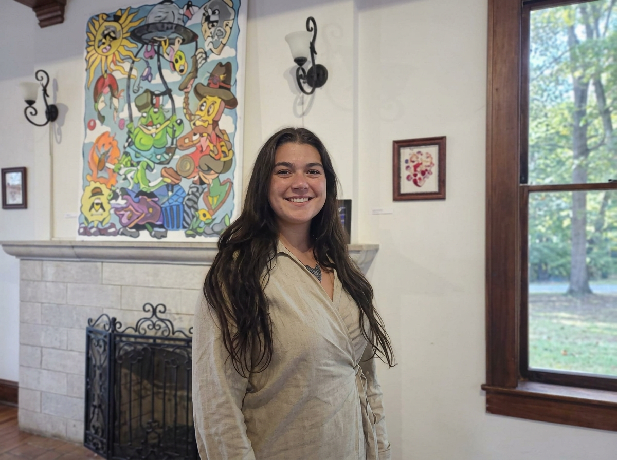 Young woman with long wavy brown hair smiling indoors, standing in front of a stone fireplace with colorful cartoon-style art above it, and a large window with wooden trim showing green trees outside.