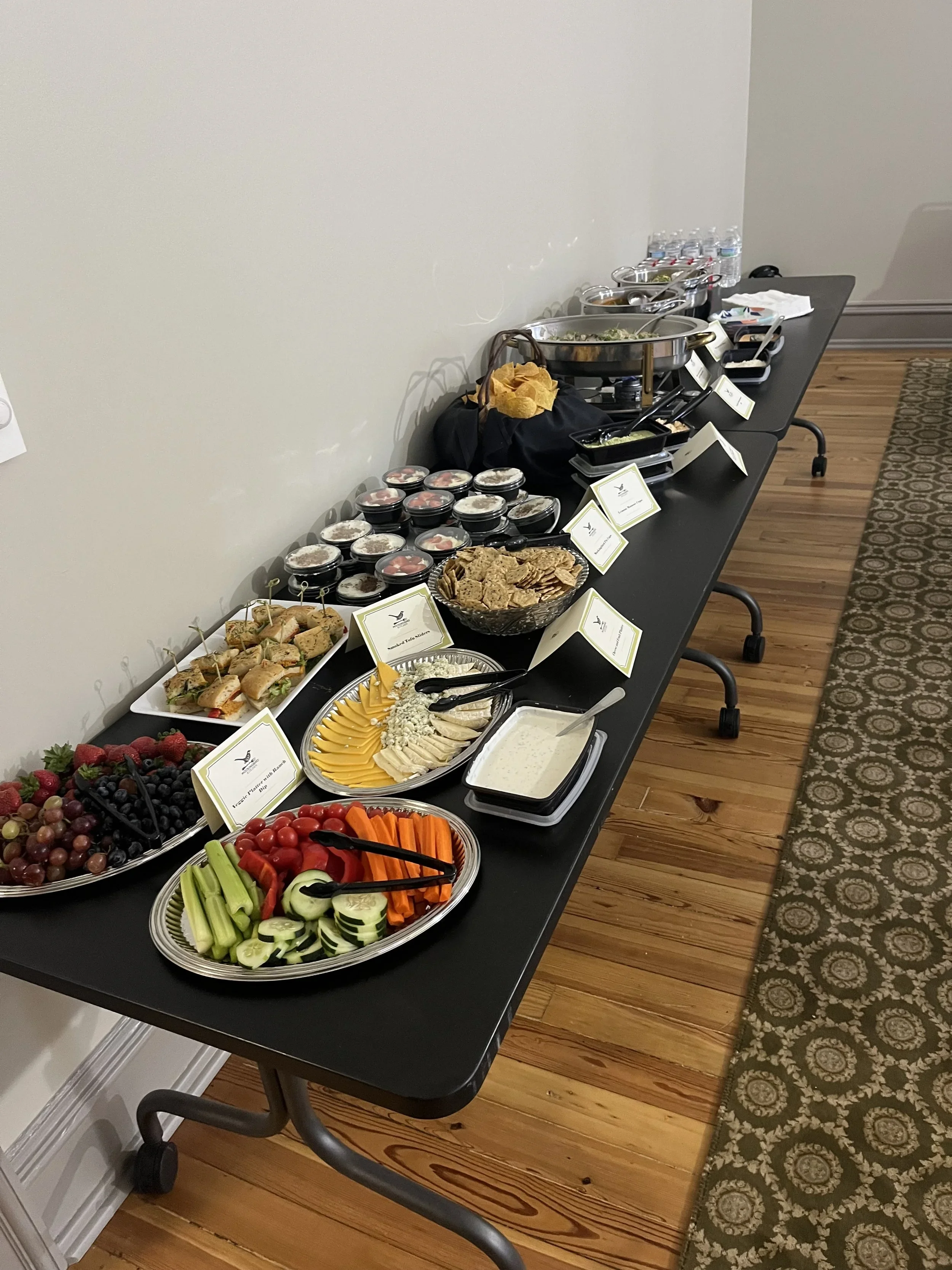 Buffet table with various salads, fruits, vegetables, crackers, cheese, and condiments. There are small labels in front of each dish, water bottles at the back, and the table is set in a room with wood flooring and patterned carpet.