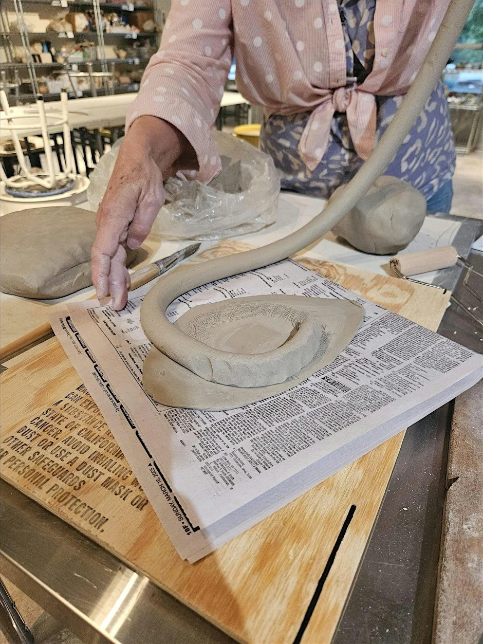 A person working on a ceramic sculpture resembling a stylized snake with a circular body, on a newspaper spread out on a wooden surface, in a pottery studio.