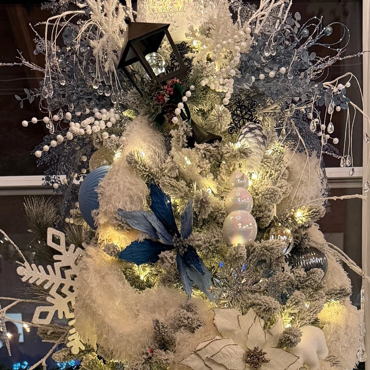 Decorated Christmas tree with white and silver ornaments, flowers, snowflakes, and a lantern, illuminated with warm lights.