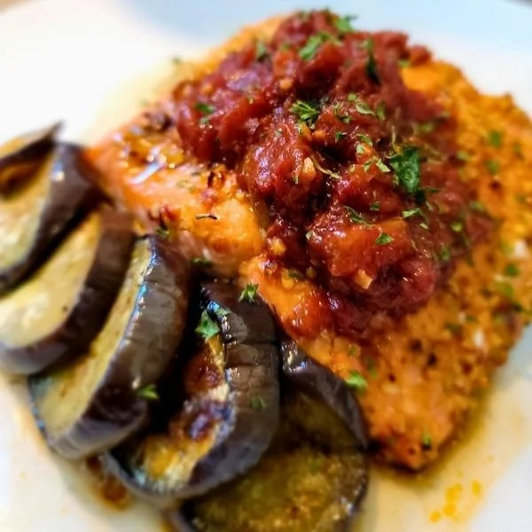 Breaded and baked fish fillet topped with tomato-based sauce and chopped herbs, served alongside roasted eggplant slices.