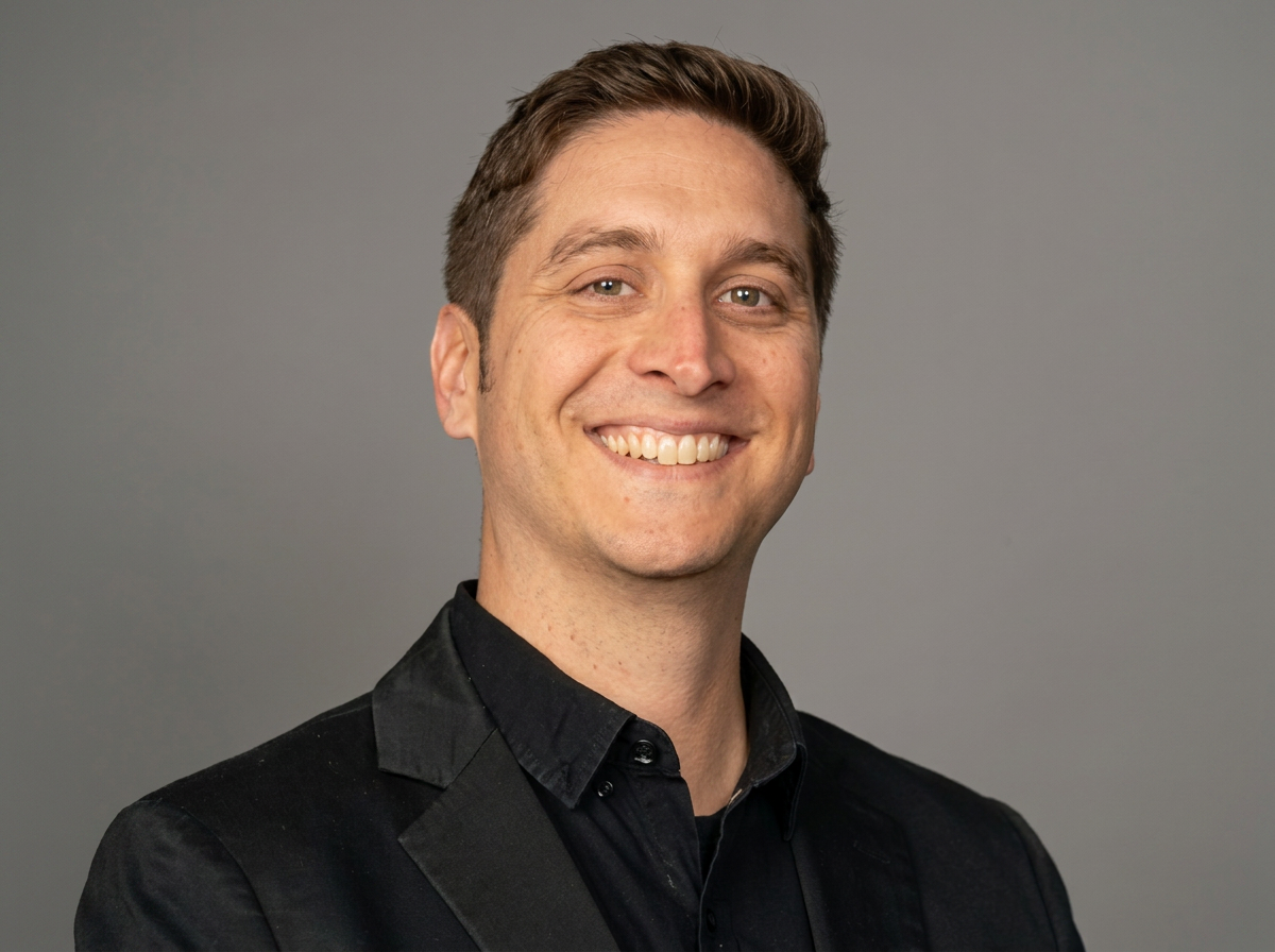 A smiling man with short brown hair and light skin, wearing a black suit jacket and shirt, against a plain gray background.