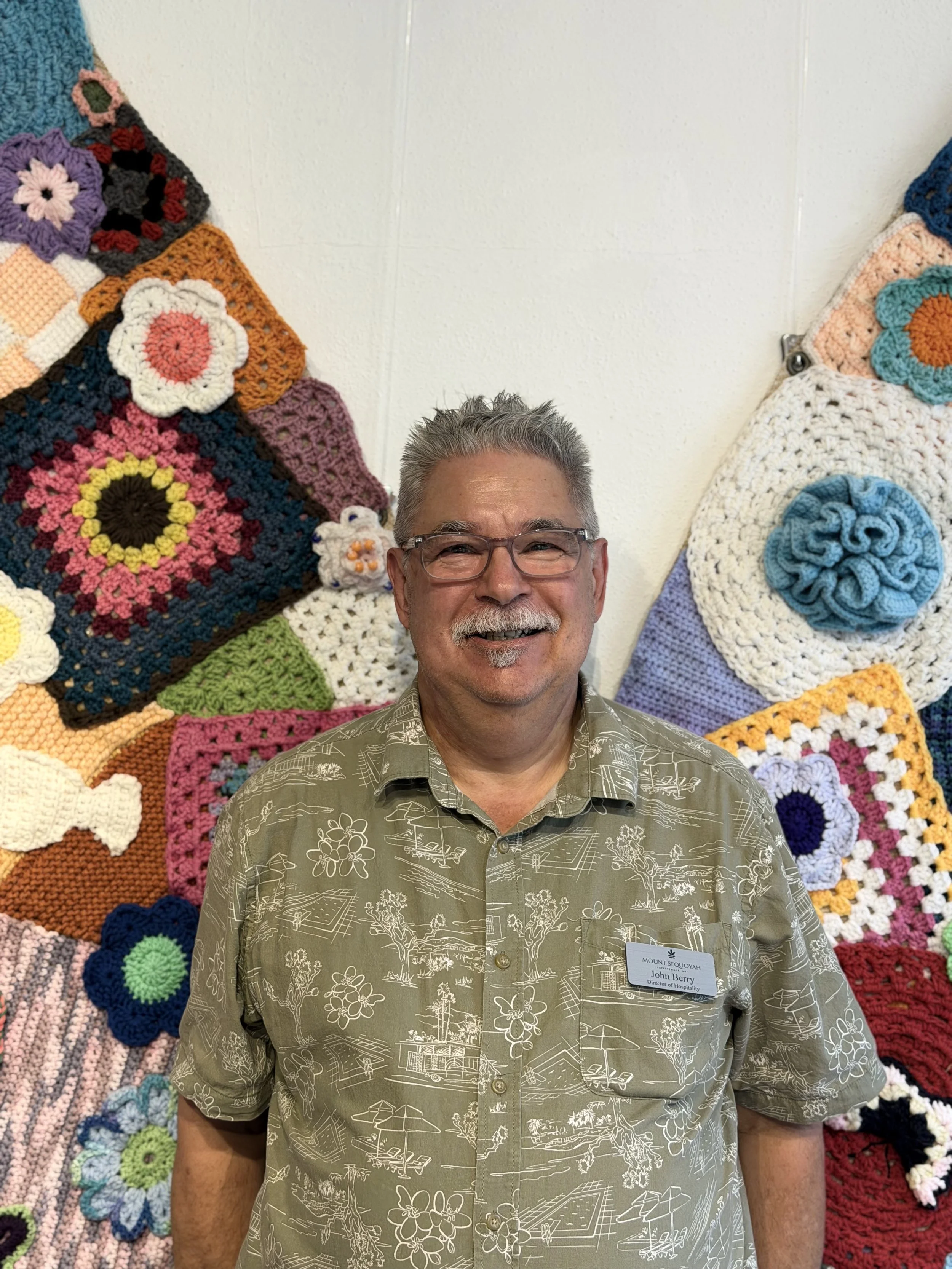 A smiling man with glasses and gray hair standing in front of a wall decorated with colorful crocheted granny squares.
