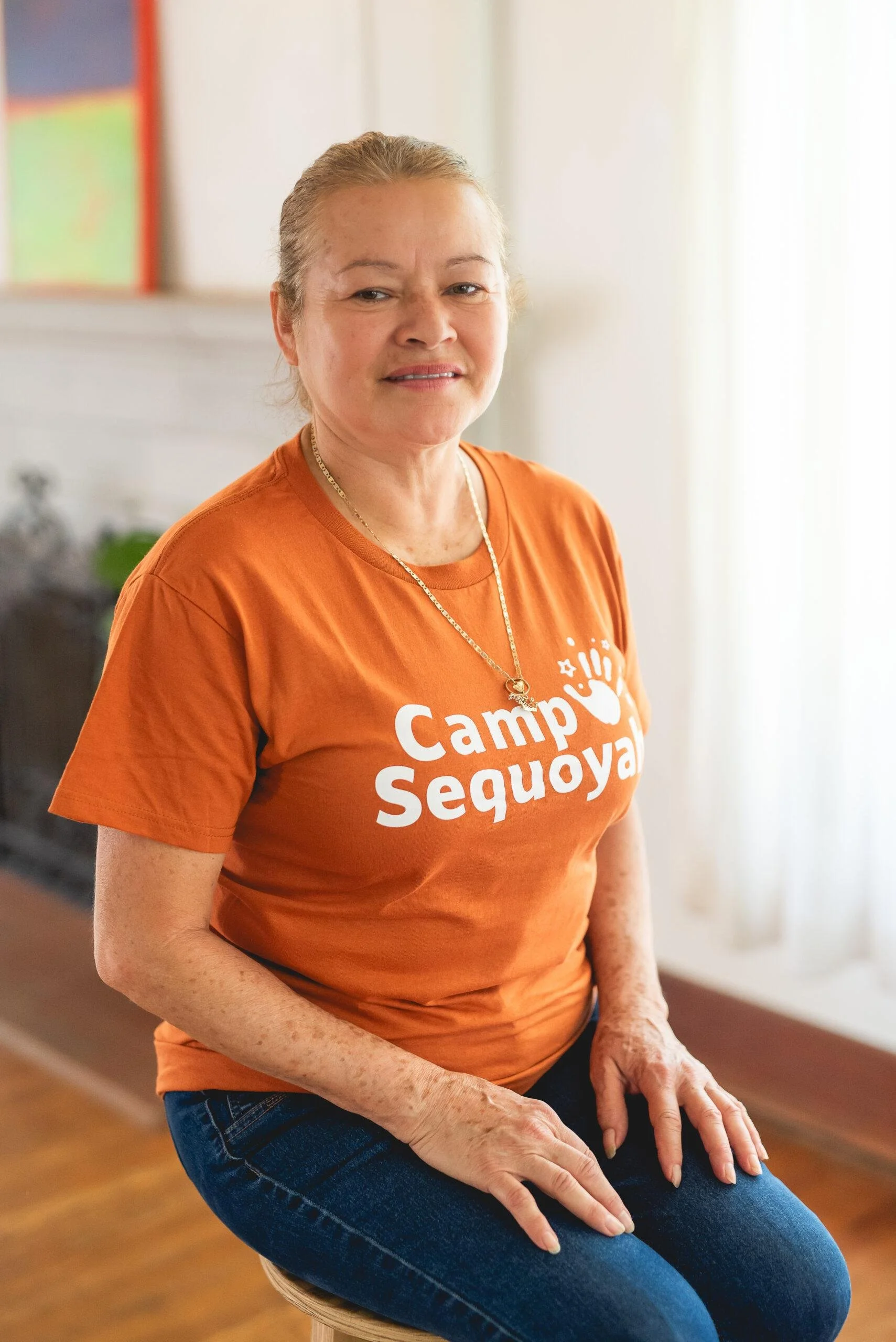 A smiling middle-aged woman with short blonde hair, wearing an orange t-shirt that says 'Camp Sequoia' and blue jeans, sitting on a stool with hands resting on her lap, indoors with a blurred background.
