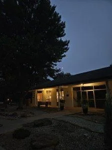 A house at dusk with outdoor lighting on, trees, and a gravel pathway in front.
