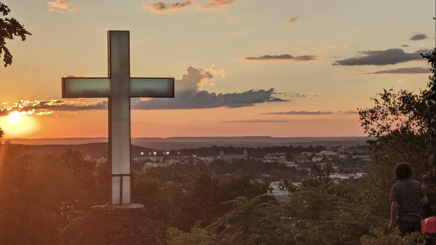 Sunset over a cityscape with a large illuminated cross in the foreground, surrounded by trees and people.