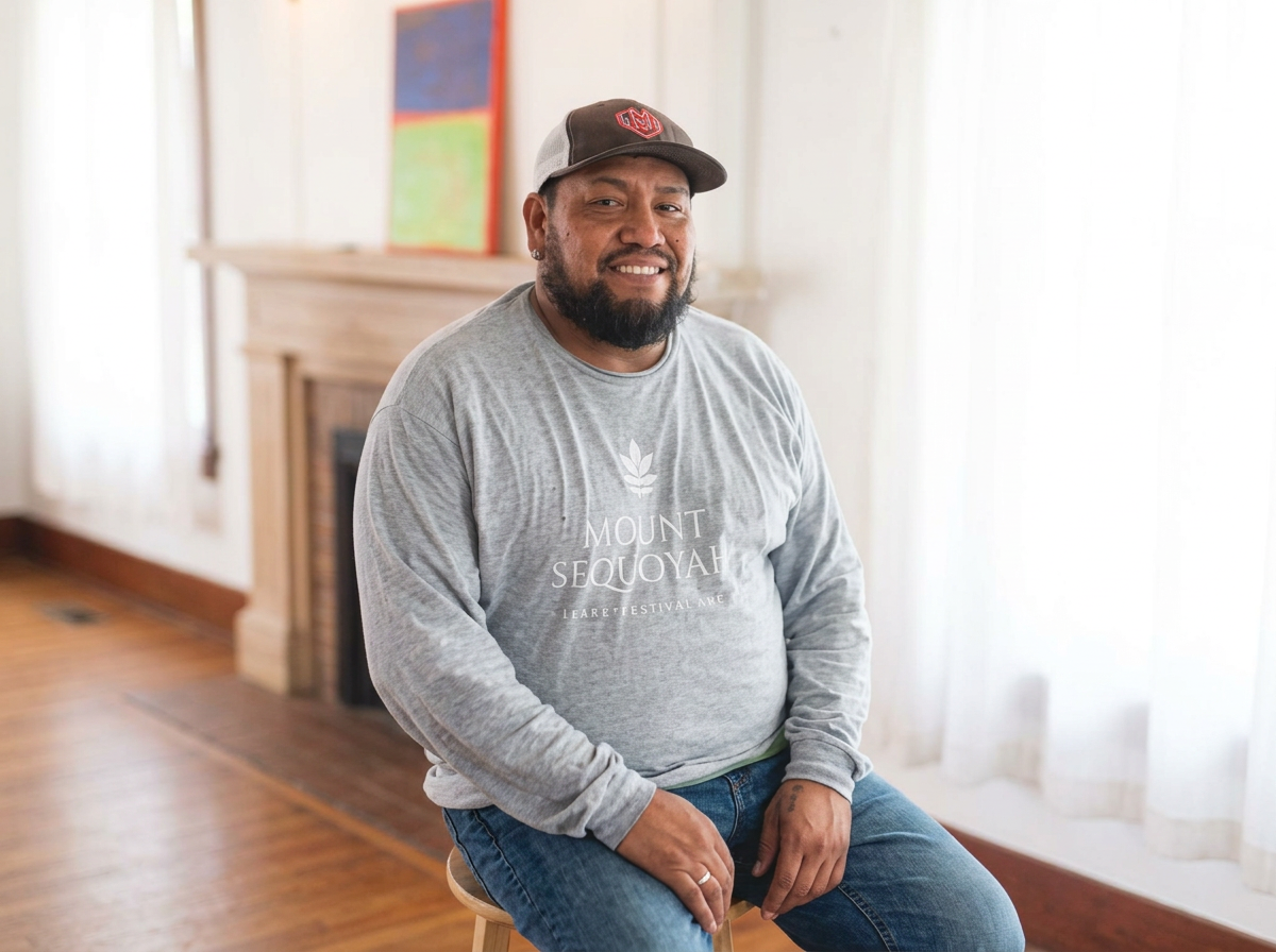 A man with a beard and a cap sitting on a chair indoors, smiling at the camera, with a fireplace and colorful artwork in the background.