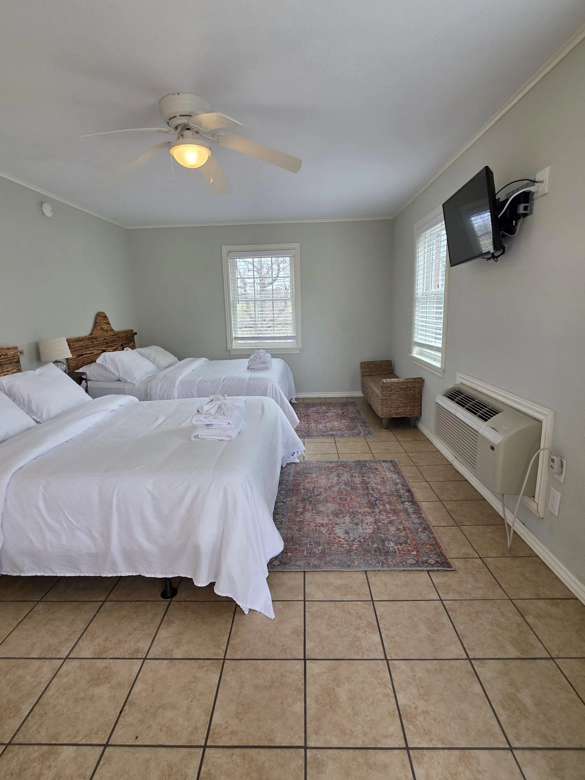 A hotel room with three beds, each with white bedding and towels, a ceiling fan with a light, two windows with blinds, a wall-mounted TV, a wicker chair, an air conditioning unit, and tiled floors with two rugs.