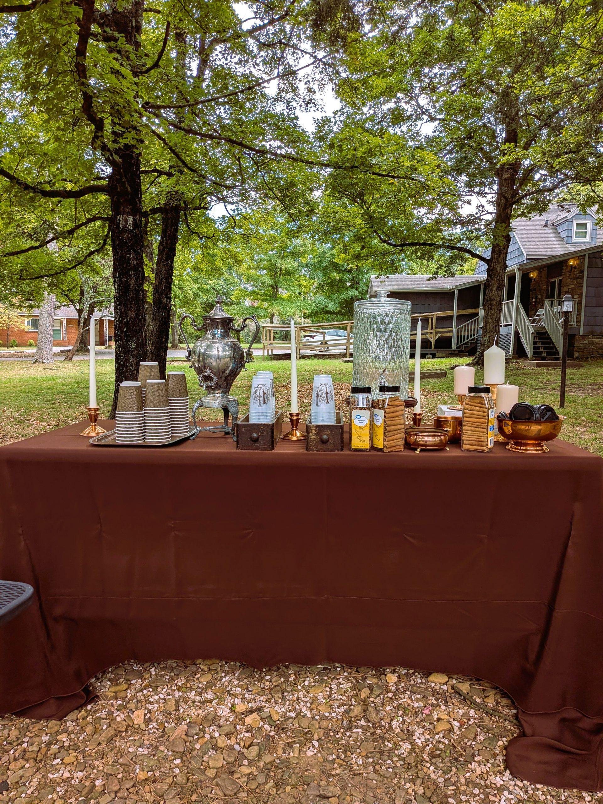 Outdoor gathering with a table covered in a brown tablecloth, featuring a large silver decorative teapot, stacks of disposable cups, and various candles, set under green trees with a house in the background.