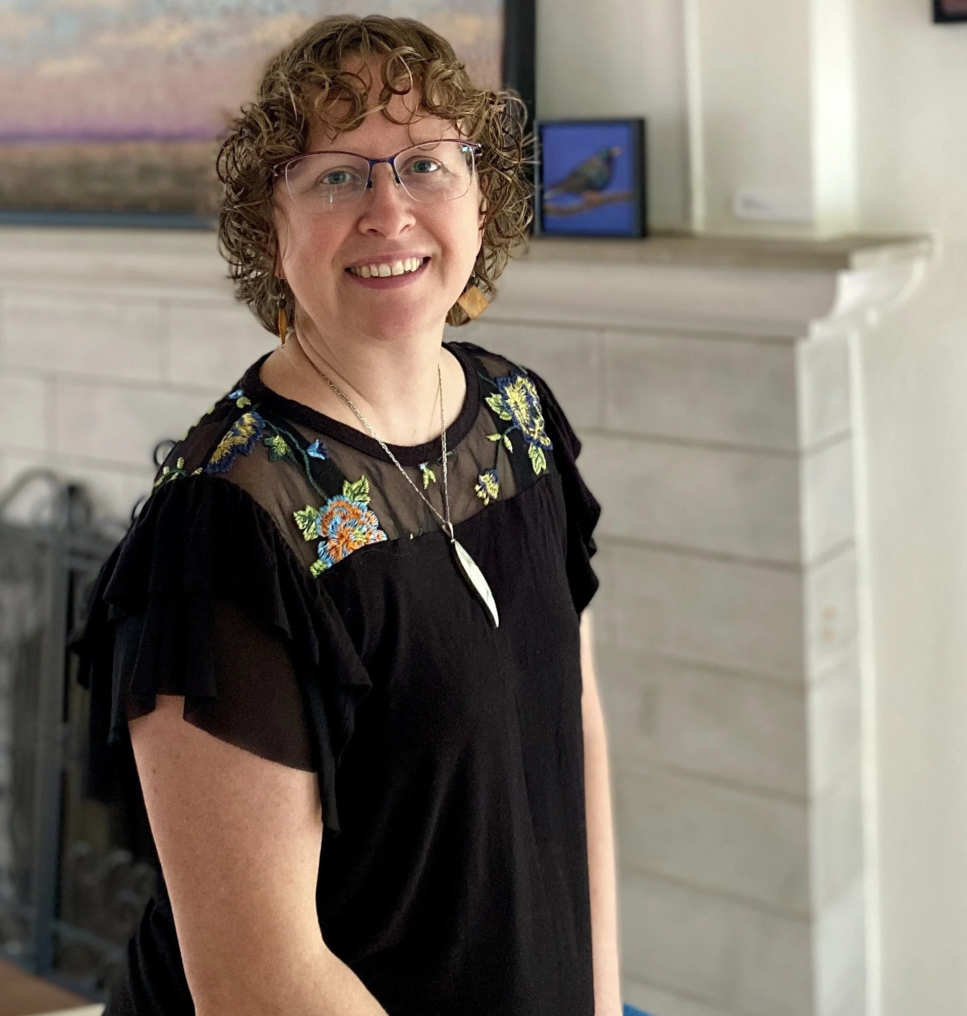 A woman with curly hair and glasses smiling indoors, wearing a black embroidered top.