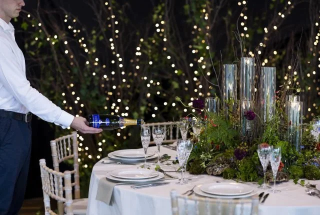 A person in a white shirt is pouring champagne into glasses at an elegant outdoor evening table with floral decorations and tall glass candle holders, illuminated by string lights in the background.