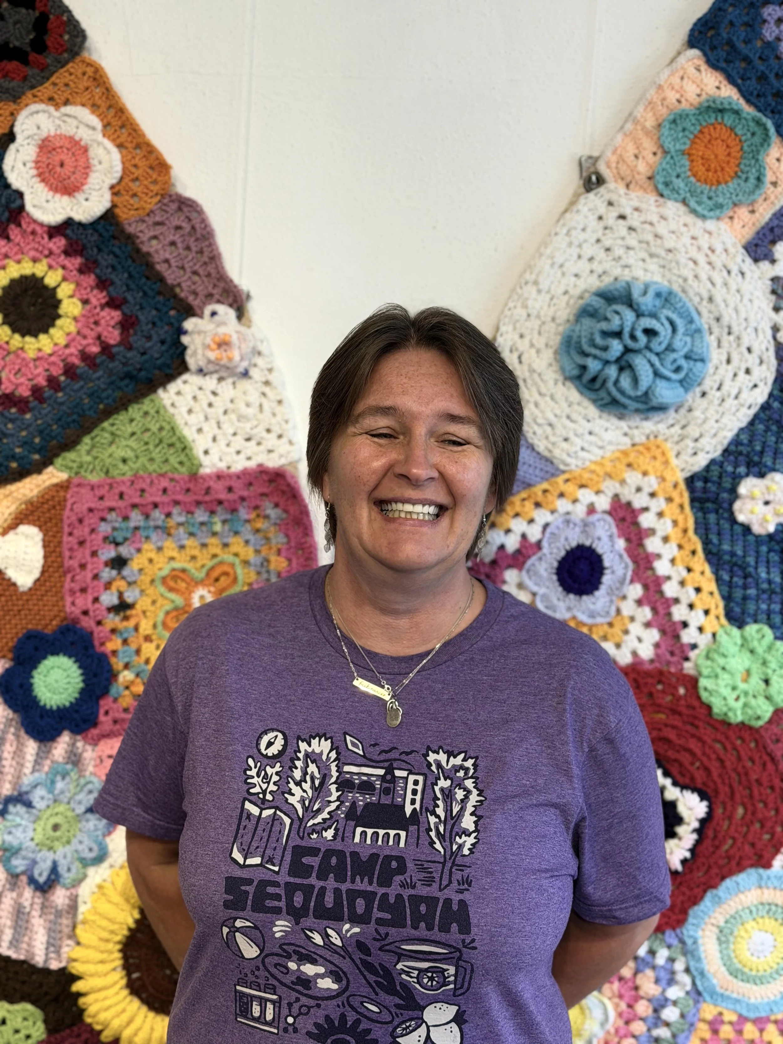 A smiling woman with short brown hair wearing a purple t-shirt with a graphic design, standing in front of a colorful crochet art wall.