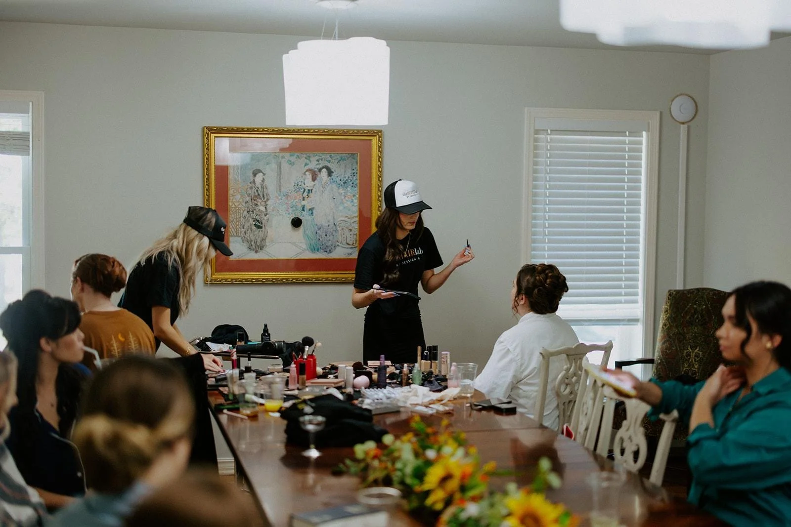 A group of women at a makeup workshop in a well-lit room with a large dining table, makeup products, and a painting on the wall. Two women are standing and demonstrating, one with a clipboard, while others are seated and observing.