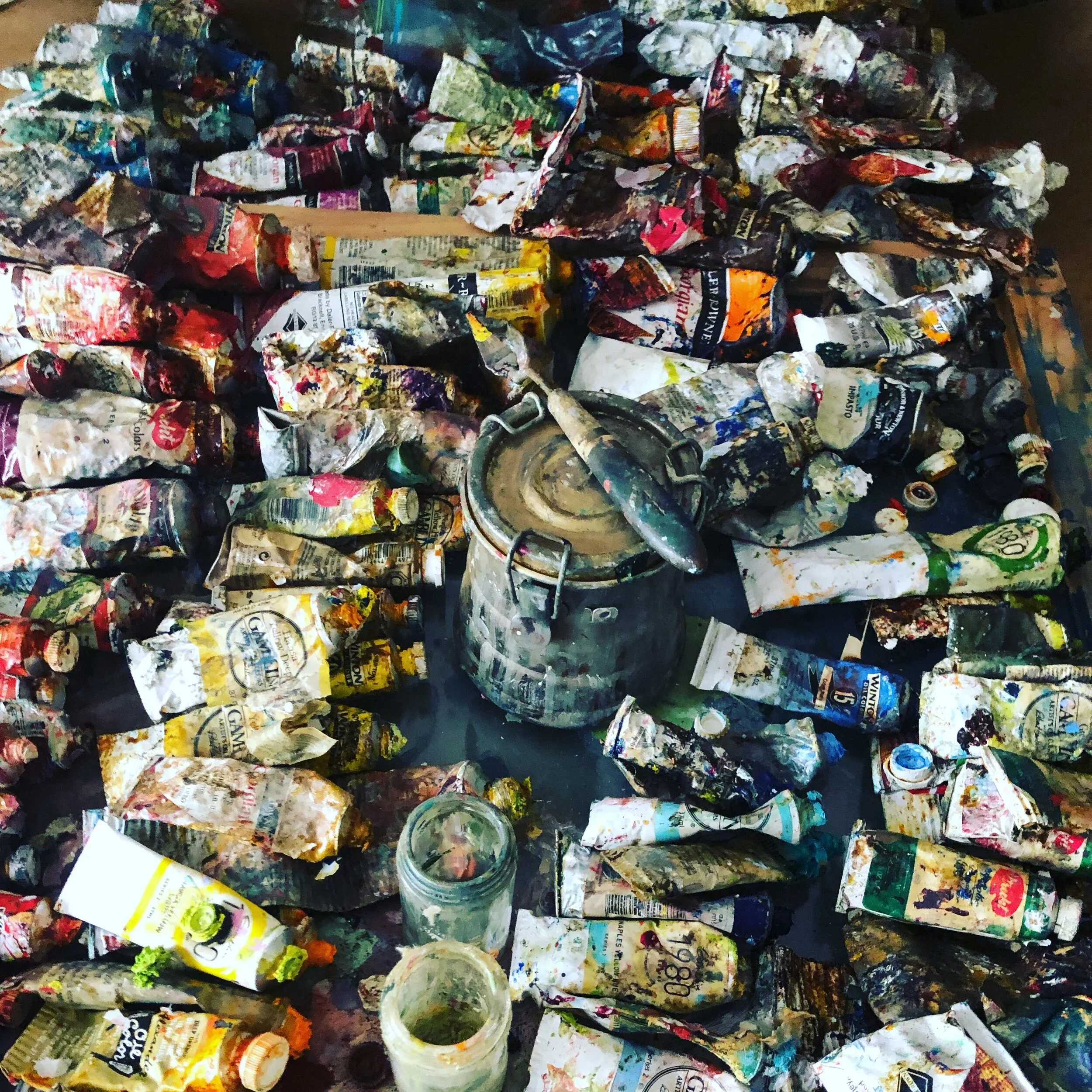 A cluttered workspace with crumpled, paint-splattered paper and used paint containers, including a jar of green paint and various paint tubes and caps scattered around.