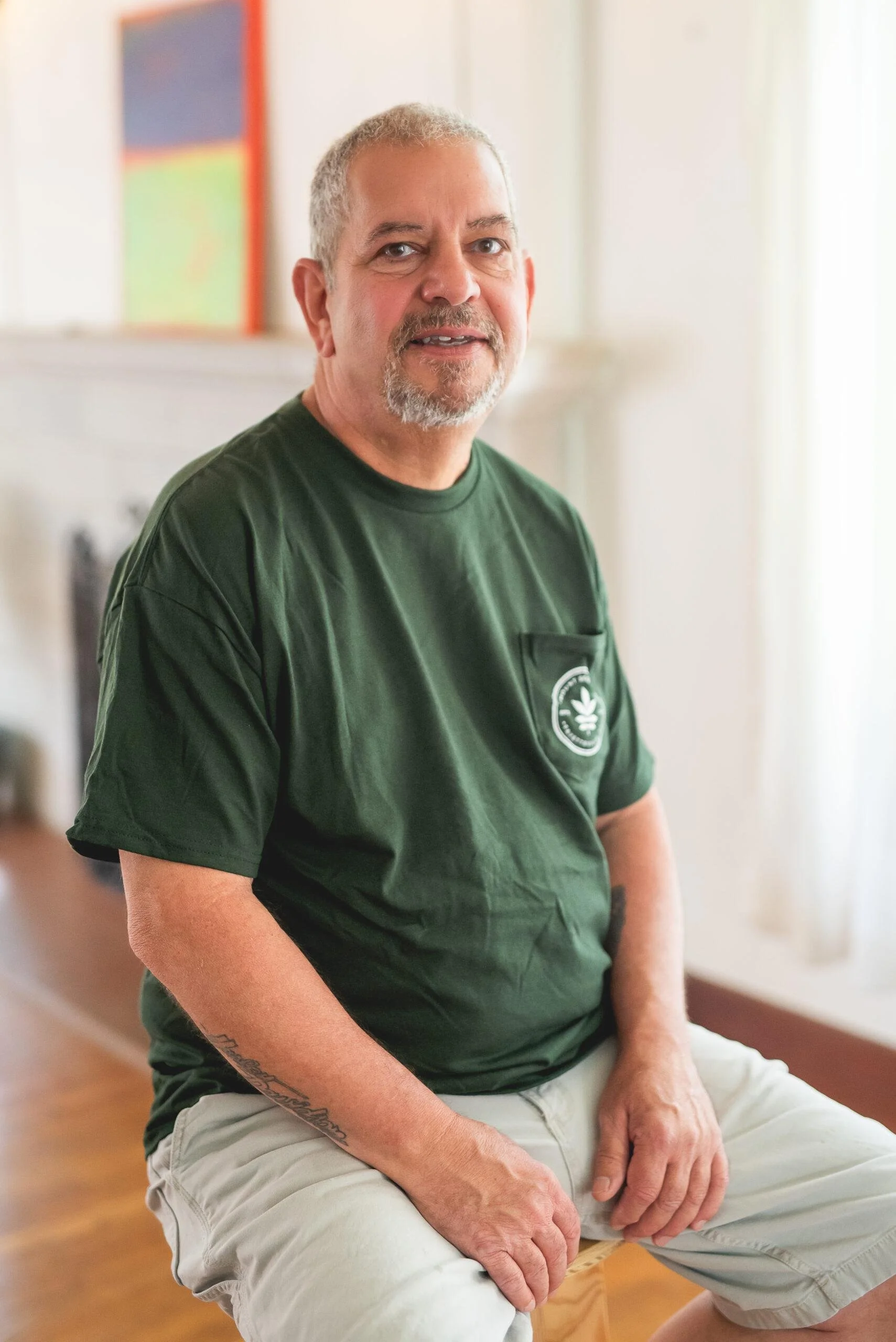 A middle-aged man with gray hair and a beard, sitting on a stool indoors. He is wearing a green T-shirt with a logo on the chest and light-colored shorts. He's looking at the camera with a slight smile.