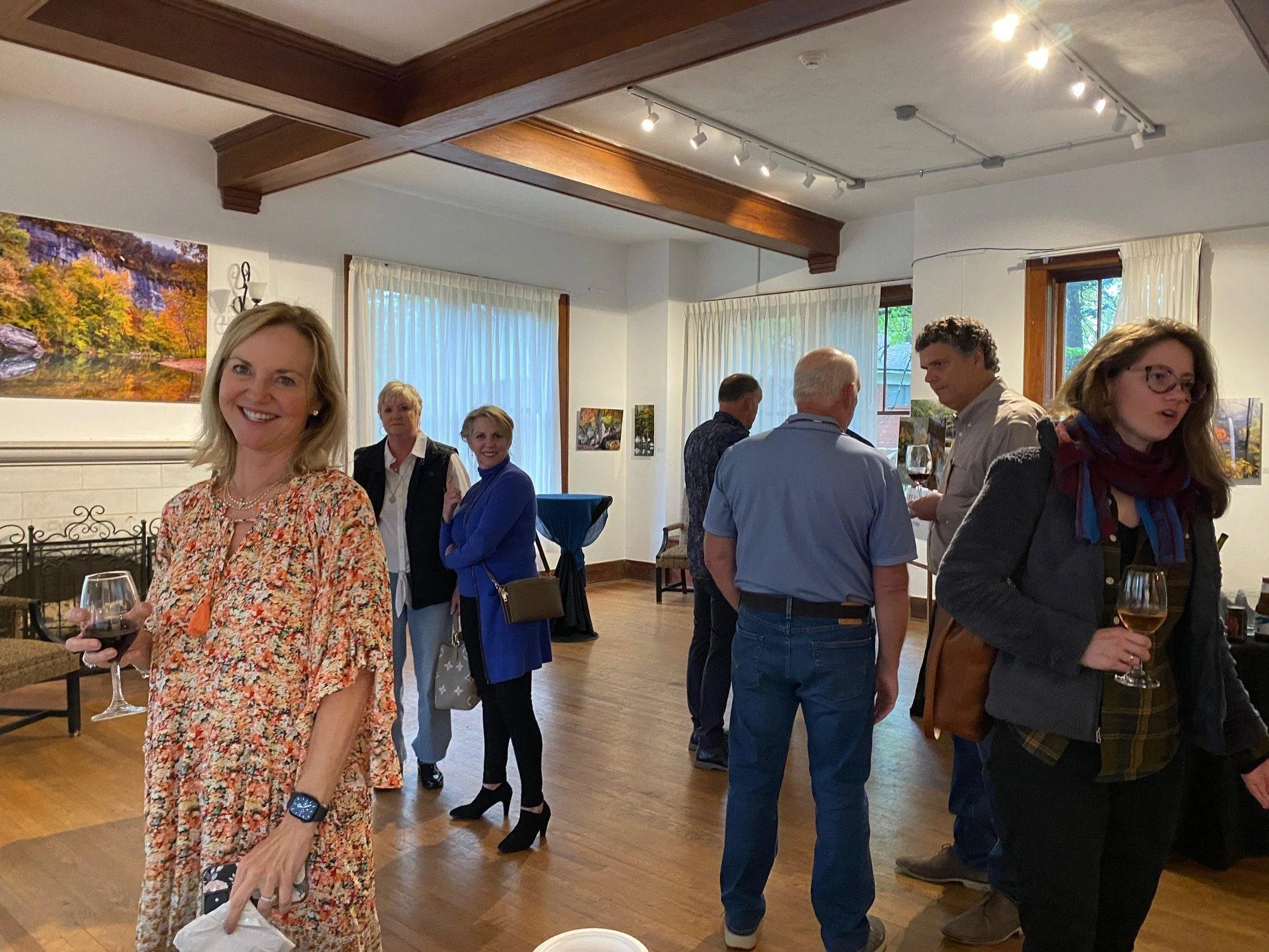 People socializing at a gathering in a room decorated with landscape paintings, some holding wine glasses, engaged in conversation.